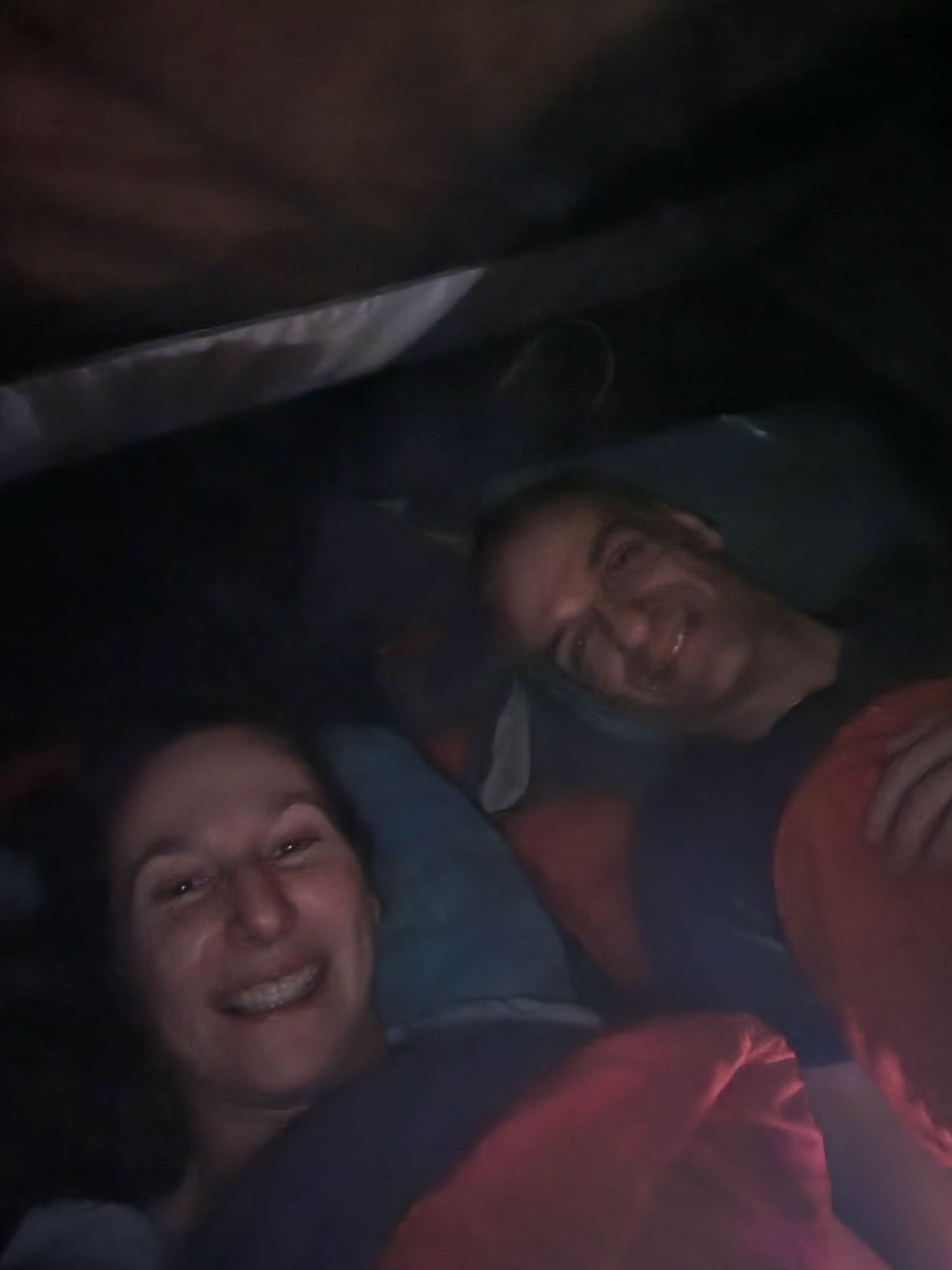 Two smiling people in sleeping bags inside a dark tent.