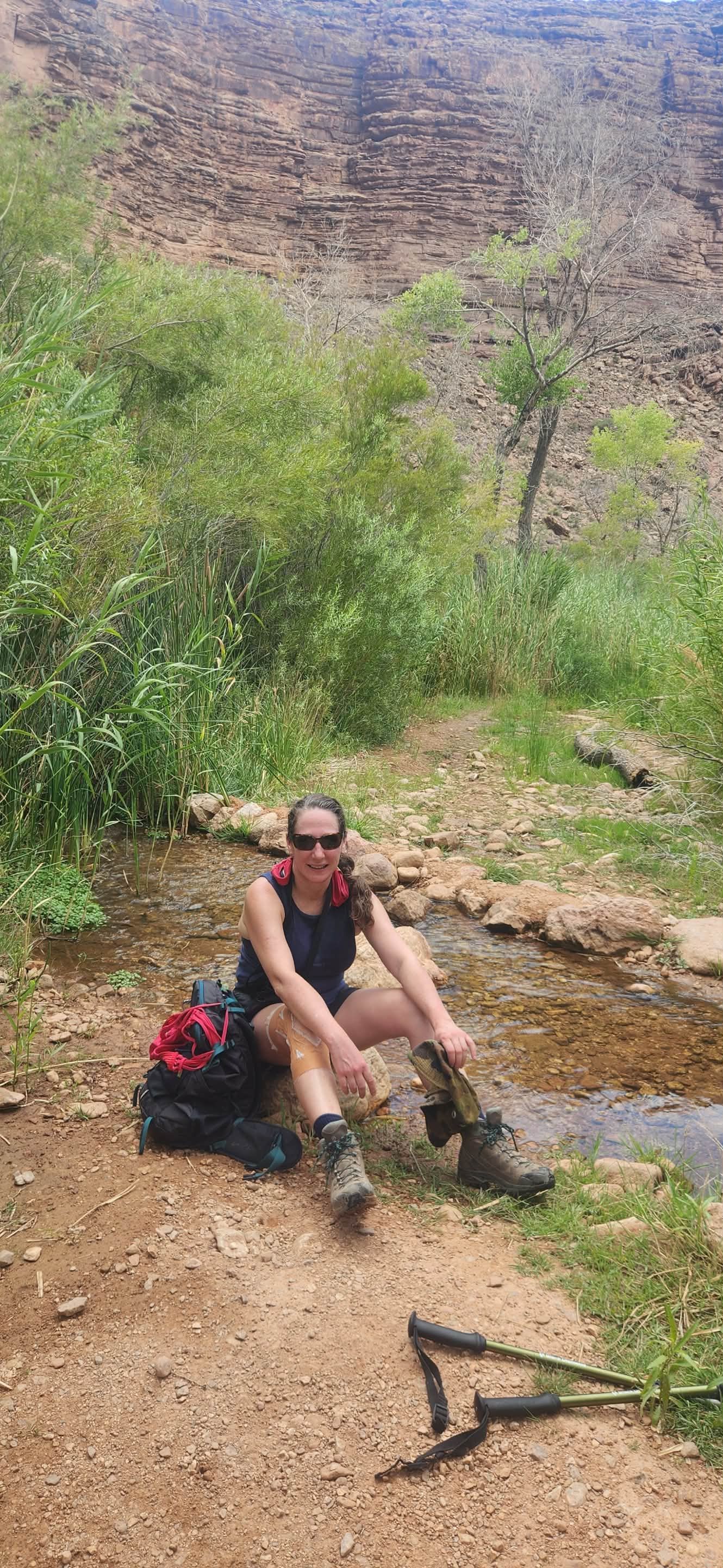 Woman hiker sitting by a stream, wearing sunglasses and hiking boots, with backpack and trekking poles.