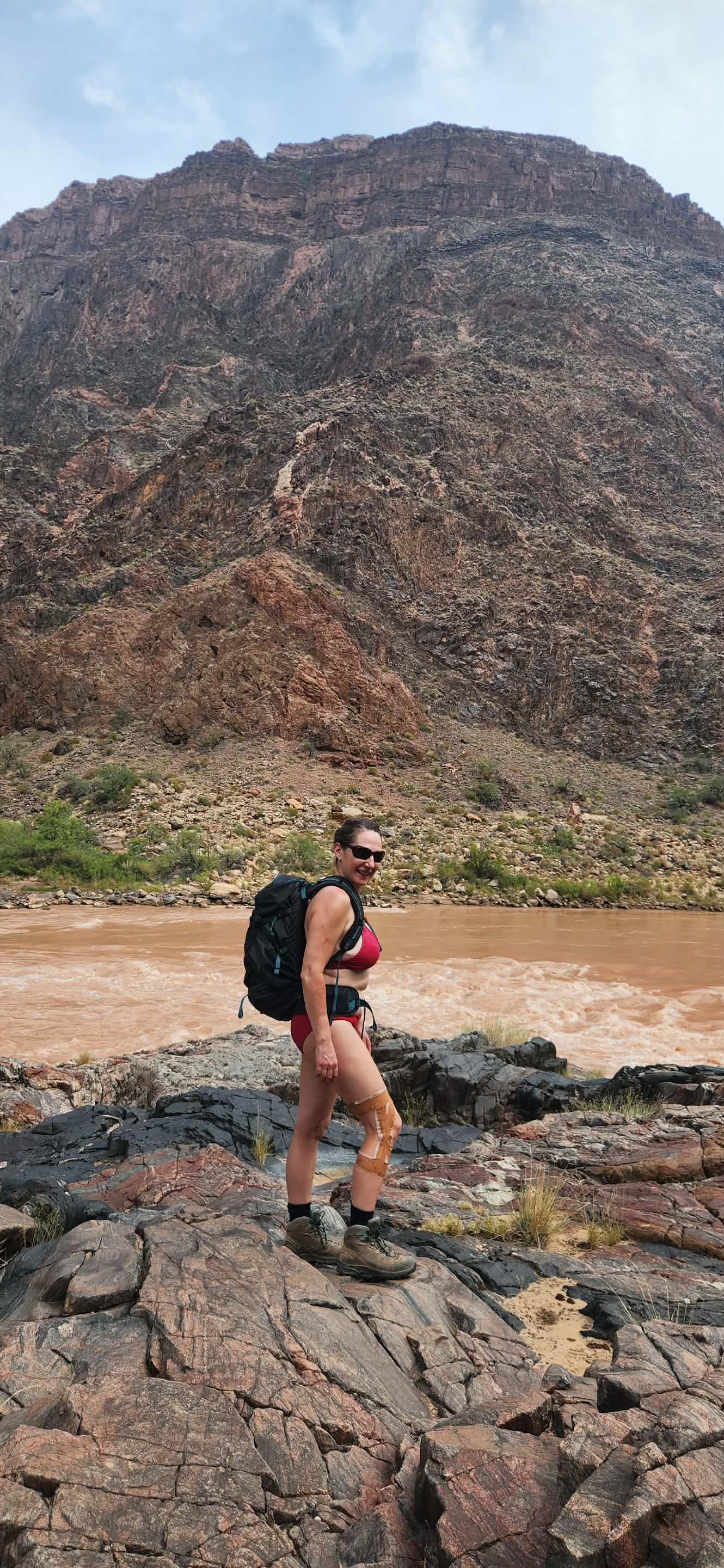 Woman with backpack stands on rocks by a river in front of a large rock formation.