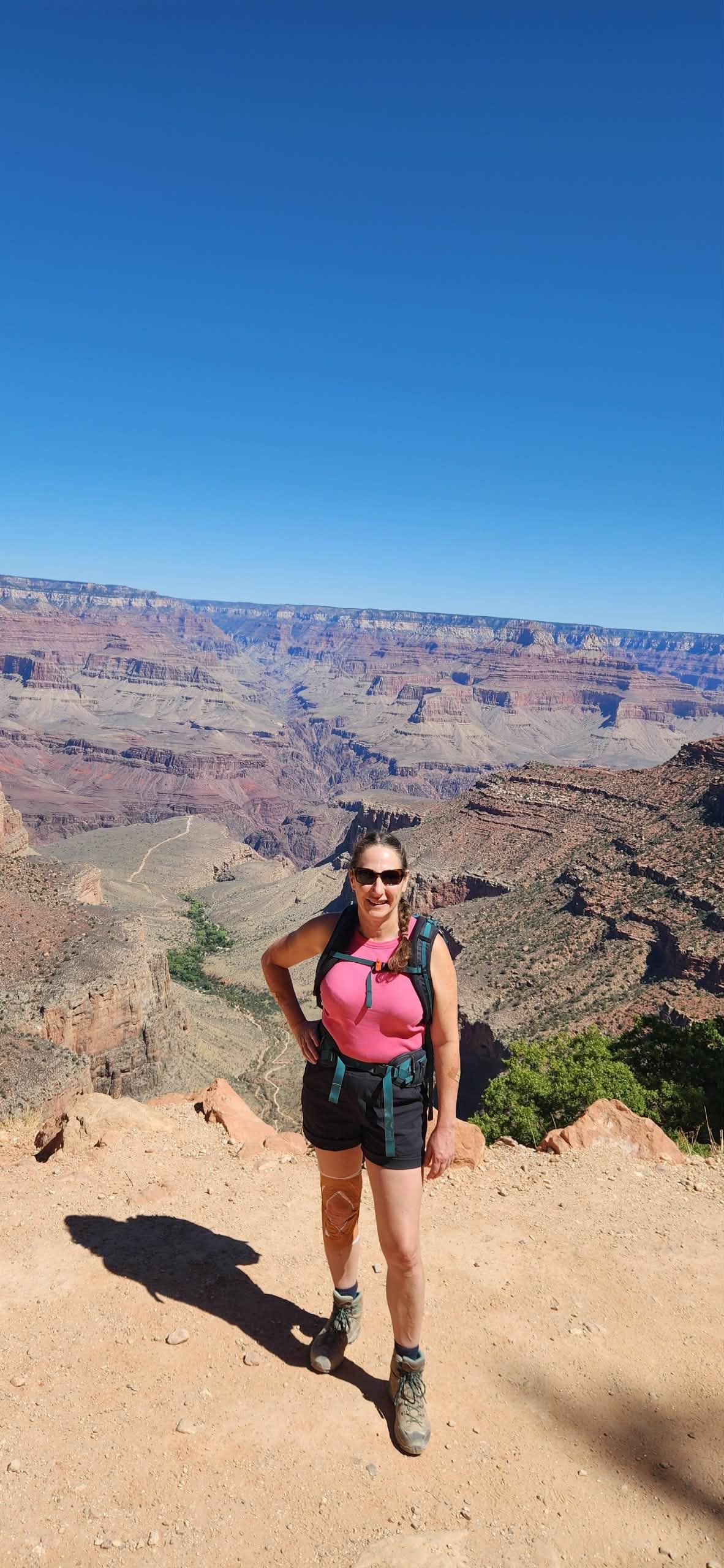 Woman posing at the Grand Canyon, wearing pink top, black shorts, hiking boots, and sunglasses.