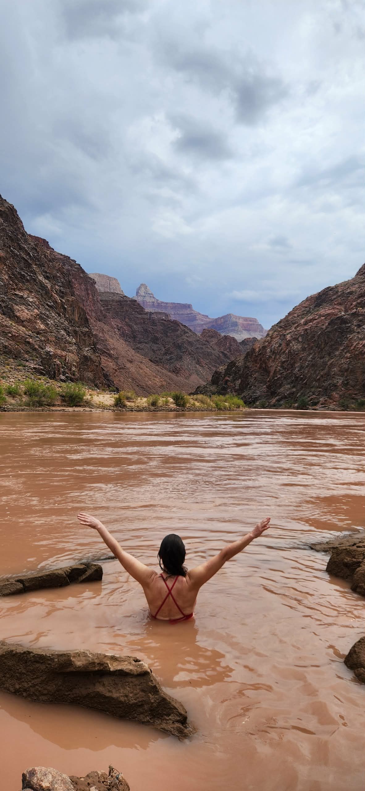Woman in water with arms raised towards canyon, cloudy sky, brown water, rocky banks.