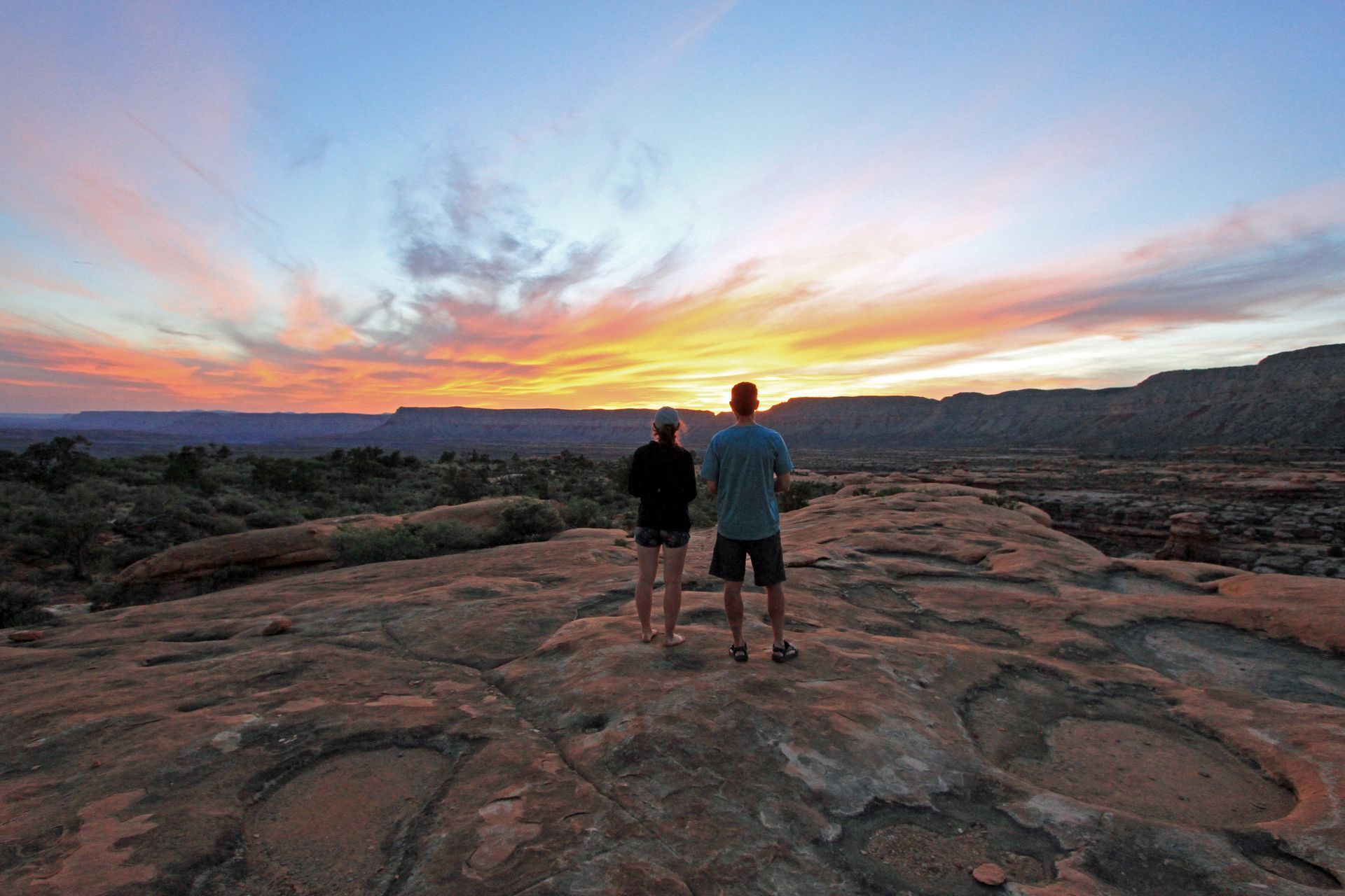 A man and a woman are standing on top of a rock watching the sunset.