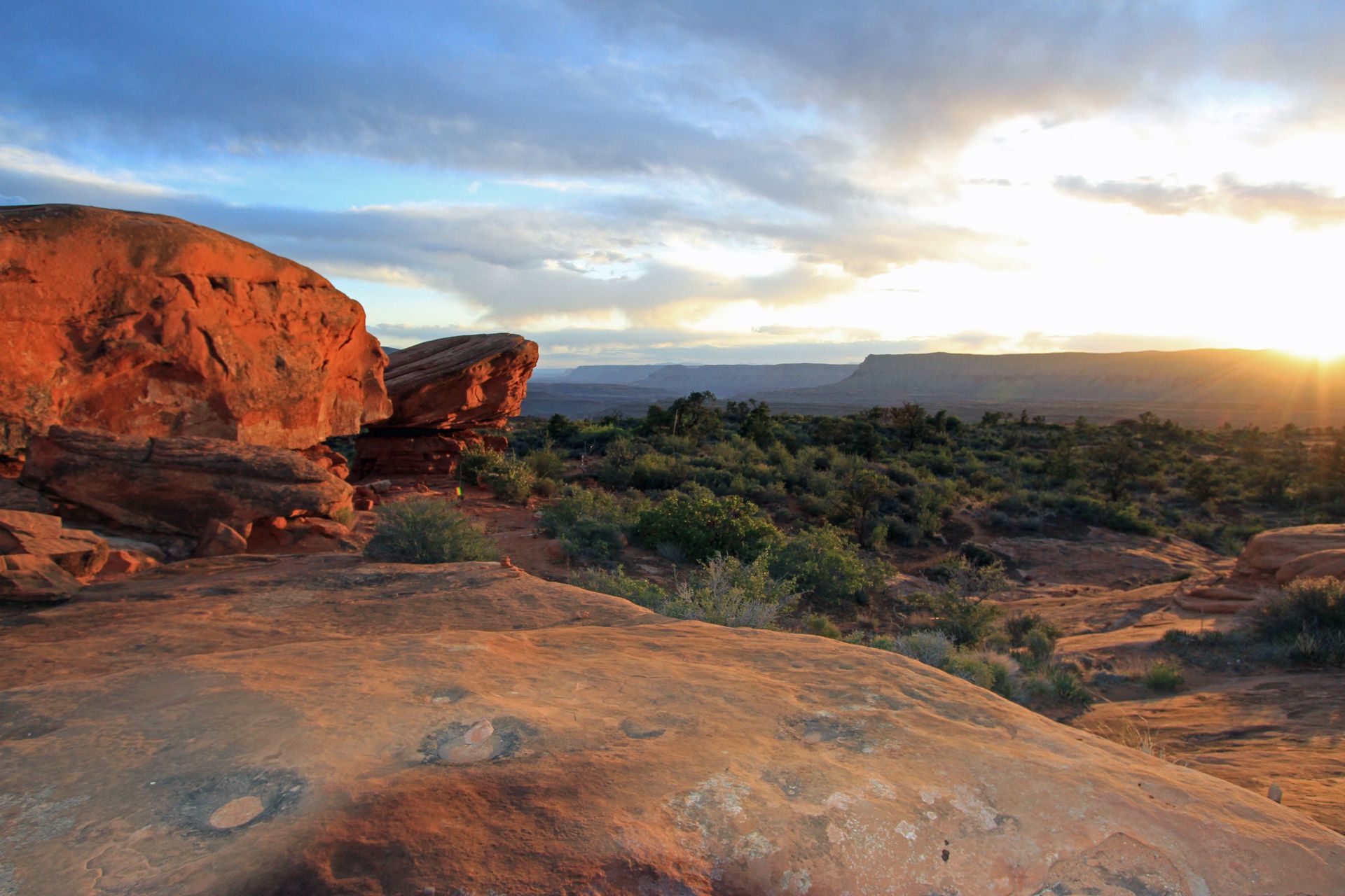 The sun is setting over a desert landscape with rocks and trees in the foreground.