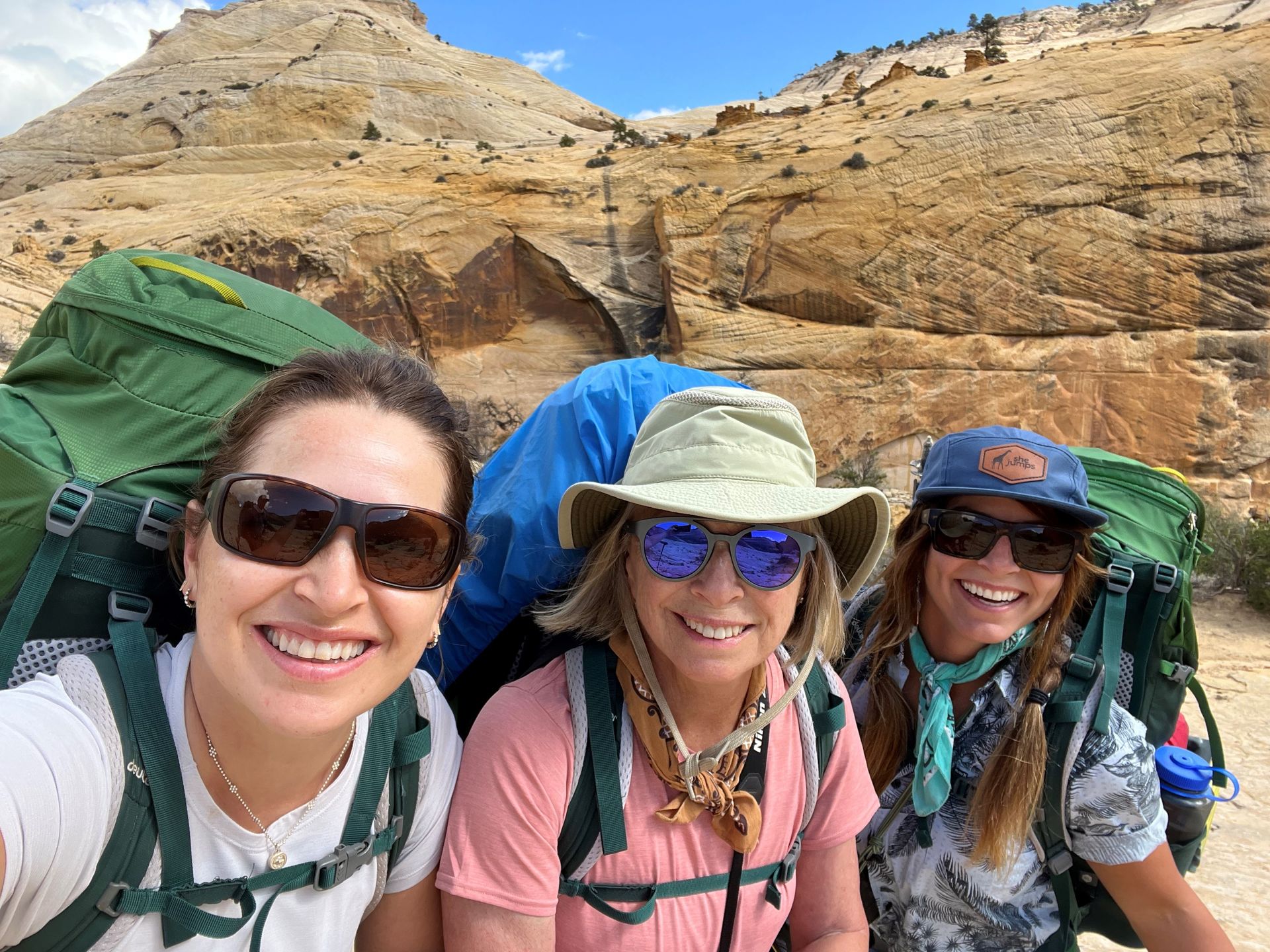 Three women wearing backpacks and sunglasses are posing for a picture.