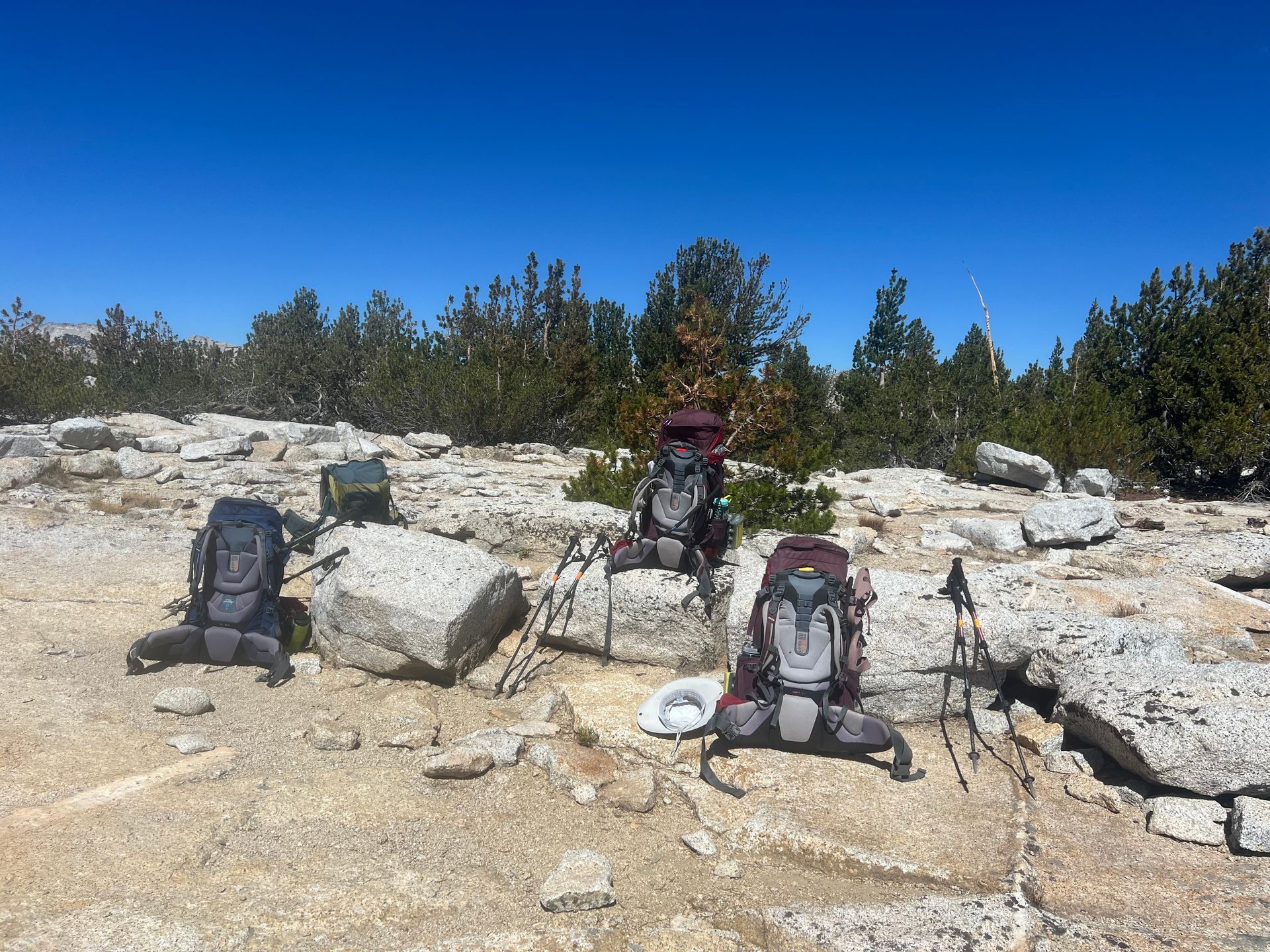 Four backpacks rest on a rocky, sunny mountaintop; hiking poles nearby.