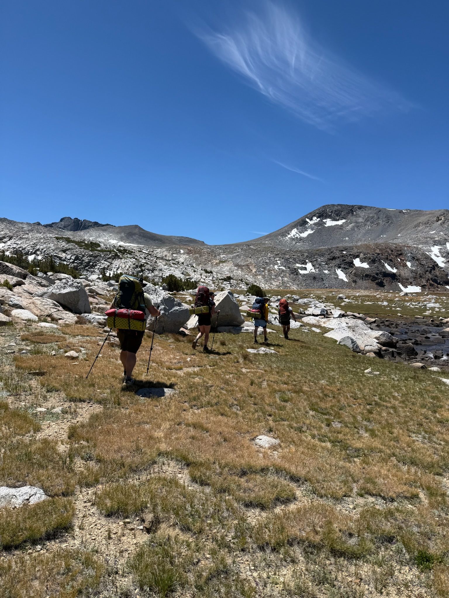 Four hikers with backpacks walking across a grassy field under a blue sky, mountains in the background.