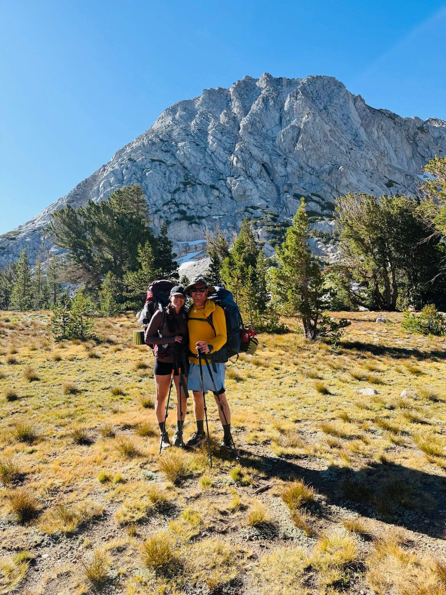Two hikers stand in a meadow with mountain backdrop. They wear backpacks and smile, sunny day.
