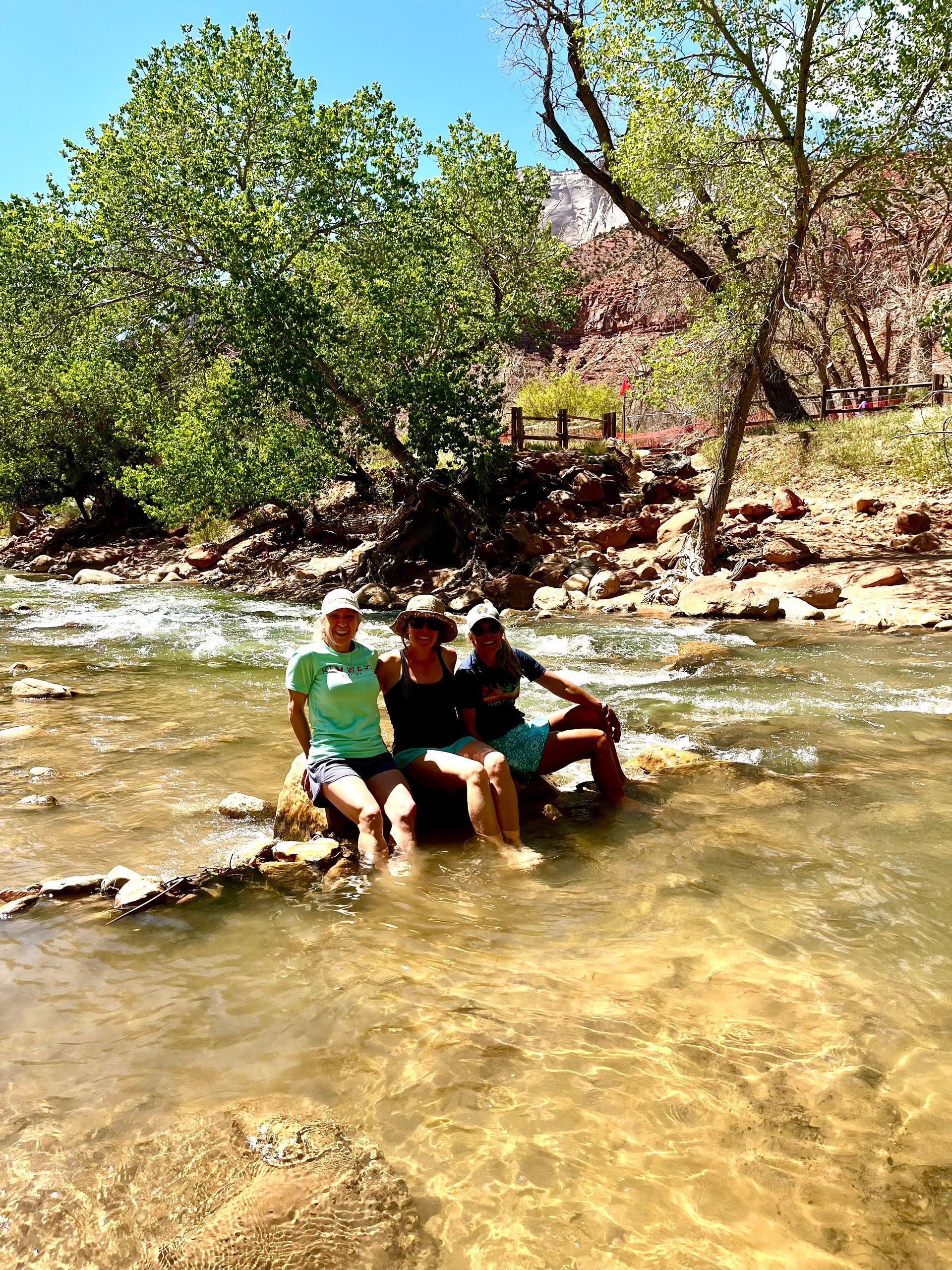 Three people sitting on rocks in a shallow, flowing river, under trees on a sunny day.