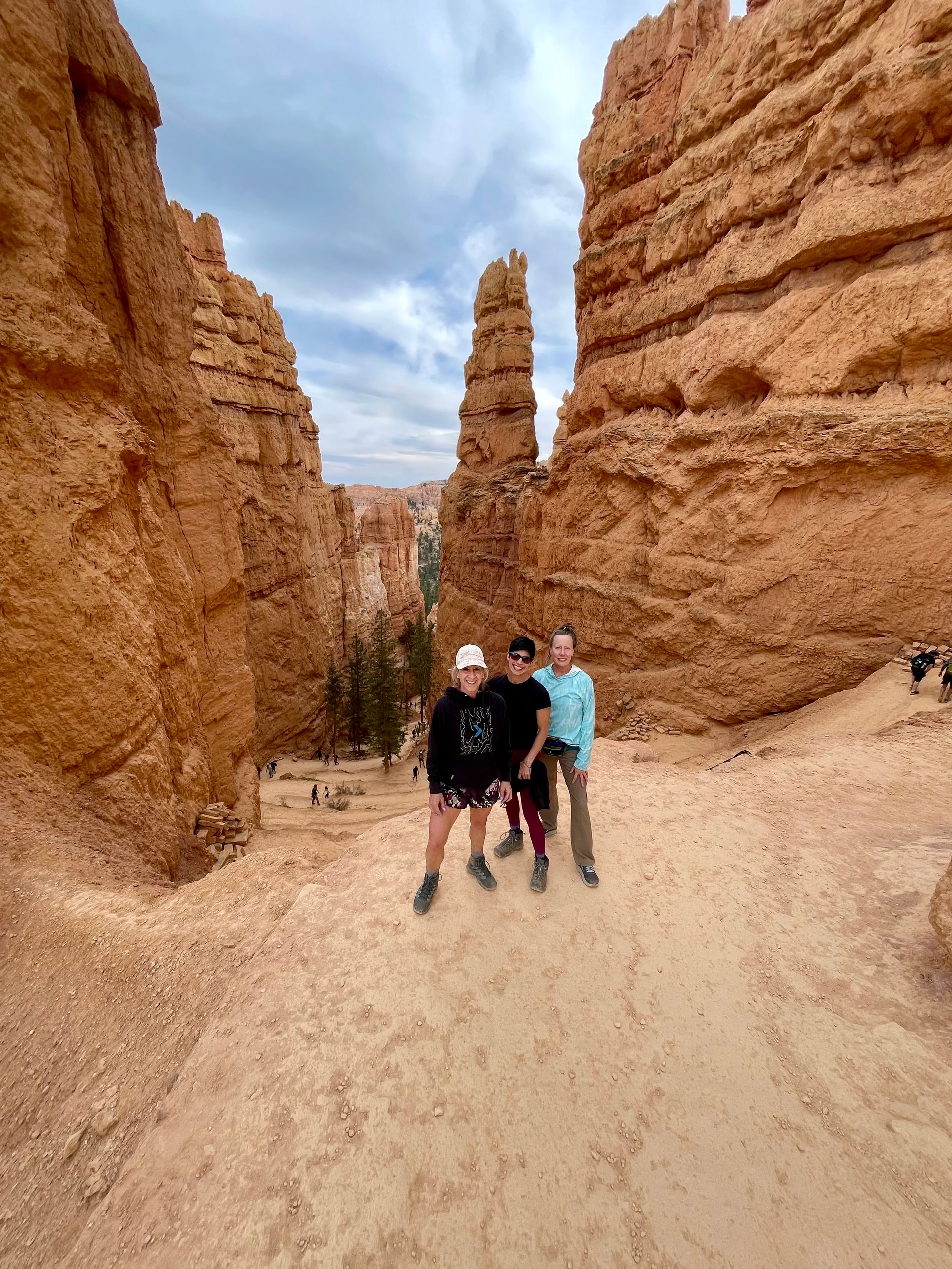 Three people stand in a narrow canyon with towering orange rock formations. Cloudy sky overhead.