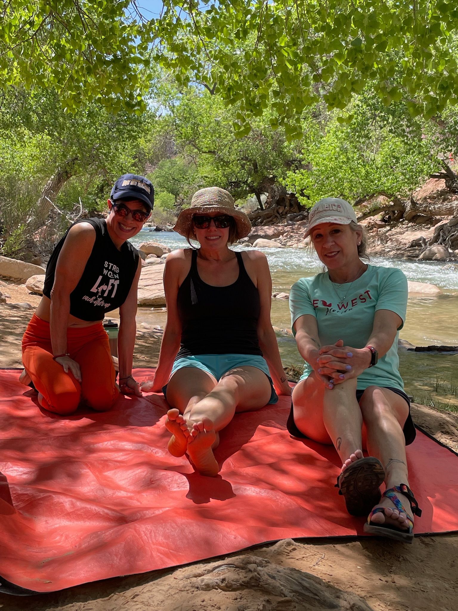 Three people sit on a red mat near a river, under tree shade. They wear athletic clothes and sunglasses.