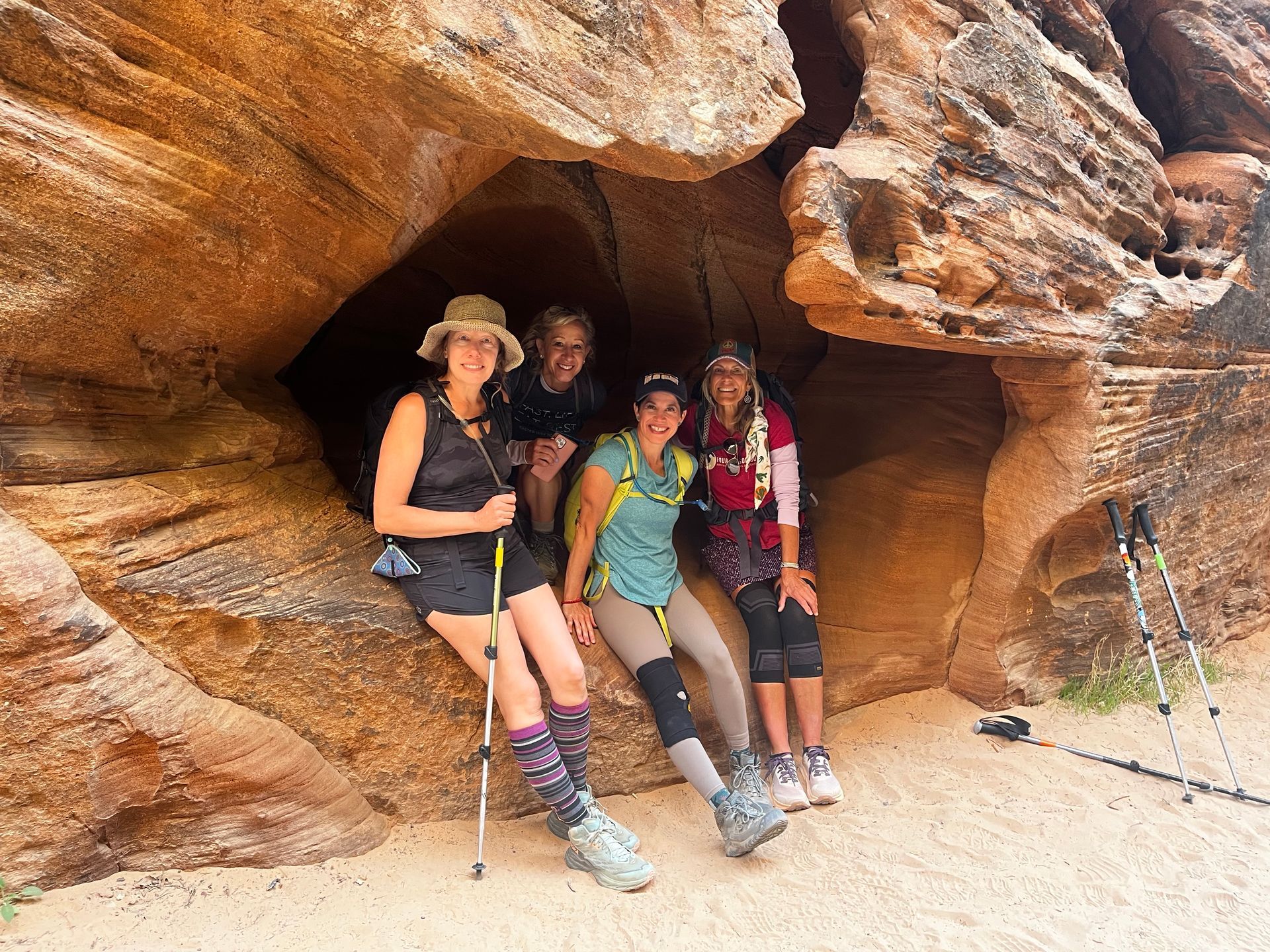Five people pose inside a sandstone cave. Smiling, they wear hiking gear. Beige and brown rock surrounds them.