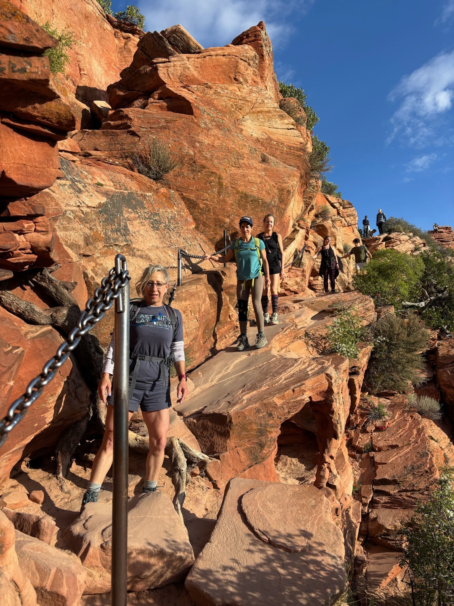 People hiking narrow trail with chains, red rock cliffs, blue sky.