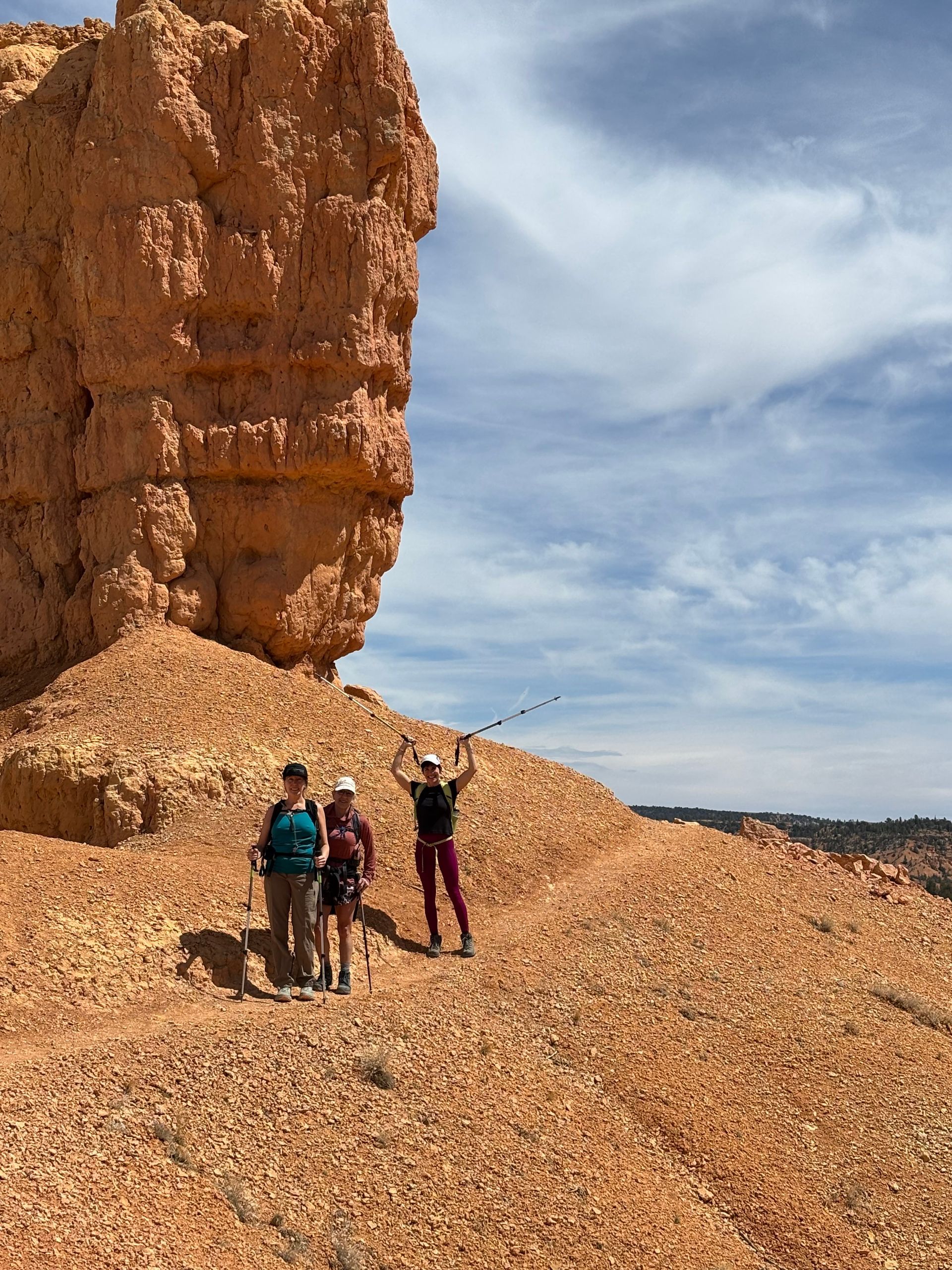 Hikers on a red-orange rocky trail, near a tall sandstone formation, under a blue sky. One raises arms.