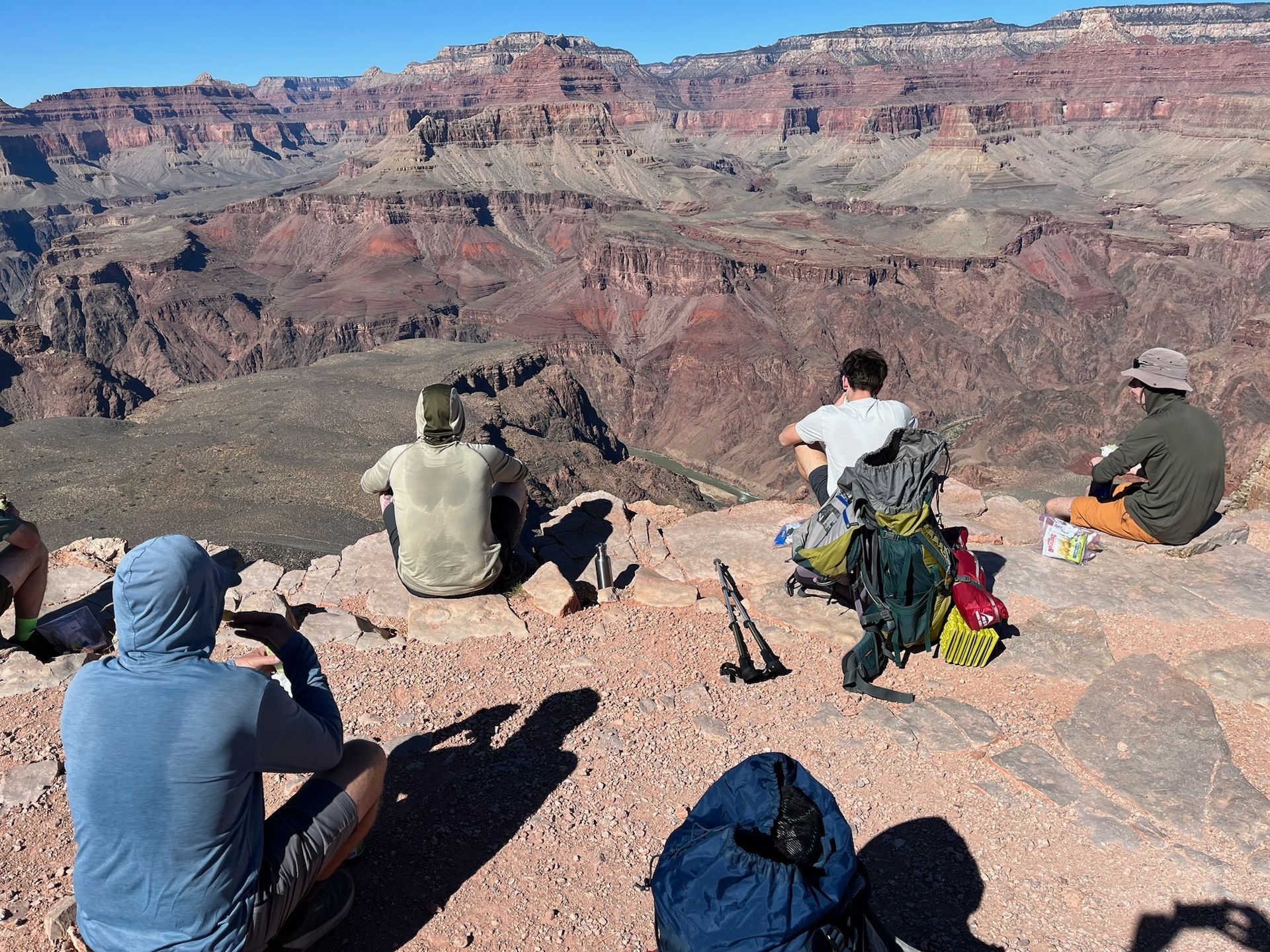 Hikers resting on a cliff, overlooking the Grand Canyon. Red rock formations dominate the background.