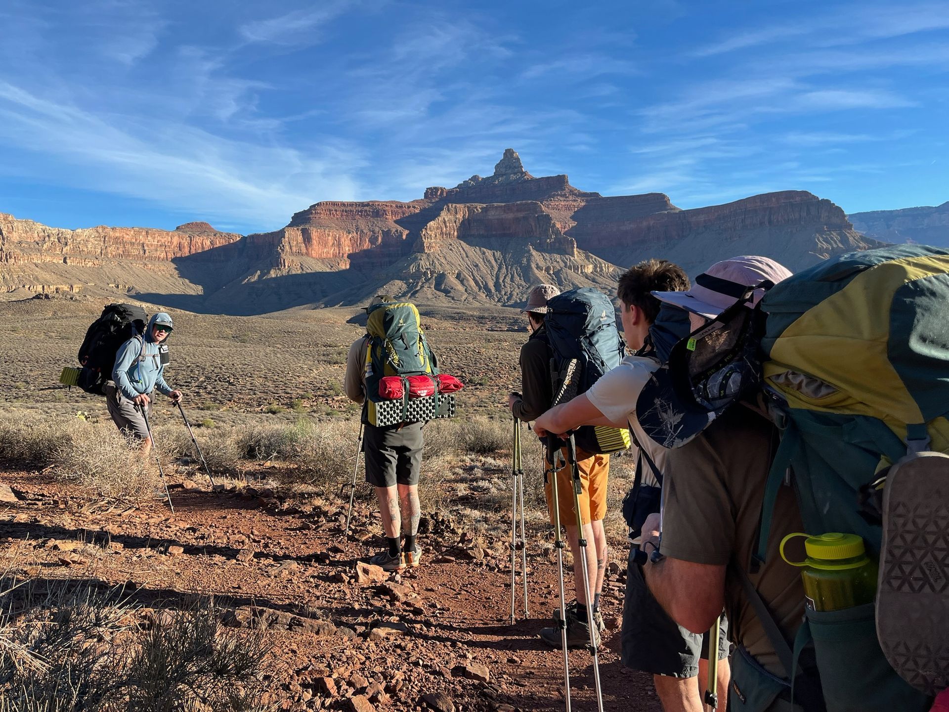 Hikers with backpacks on a trail, heading toward a large rock formation in a desert landscape.