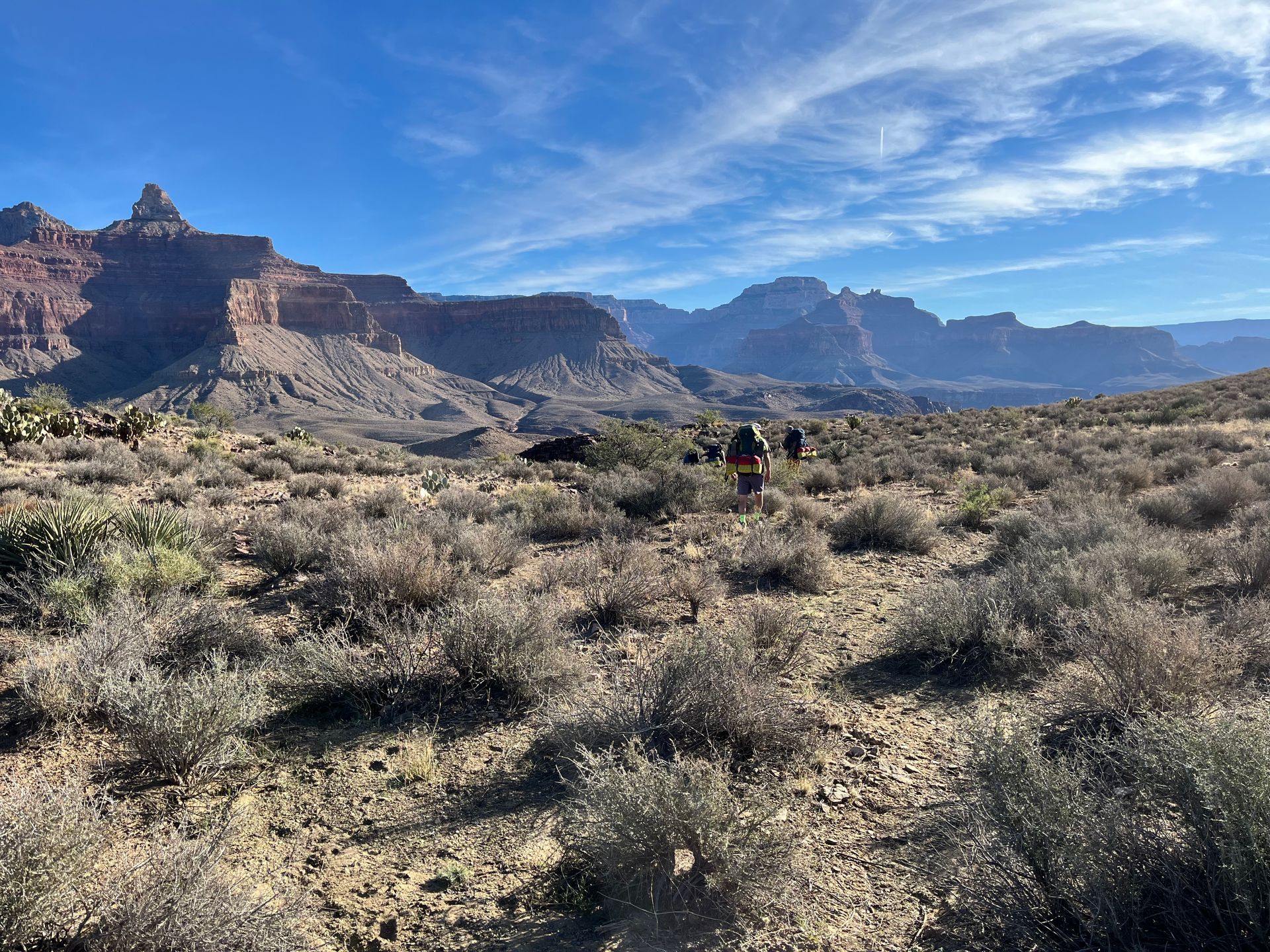 Hikers in a desert landscape with red rock formations and a blue sky dotted with clouds.
