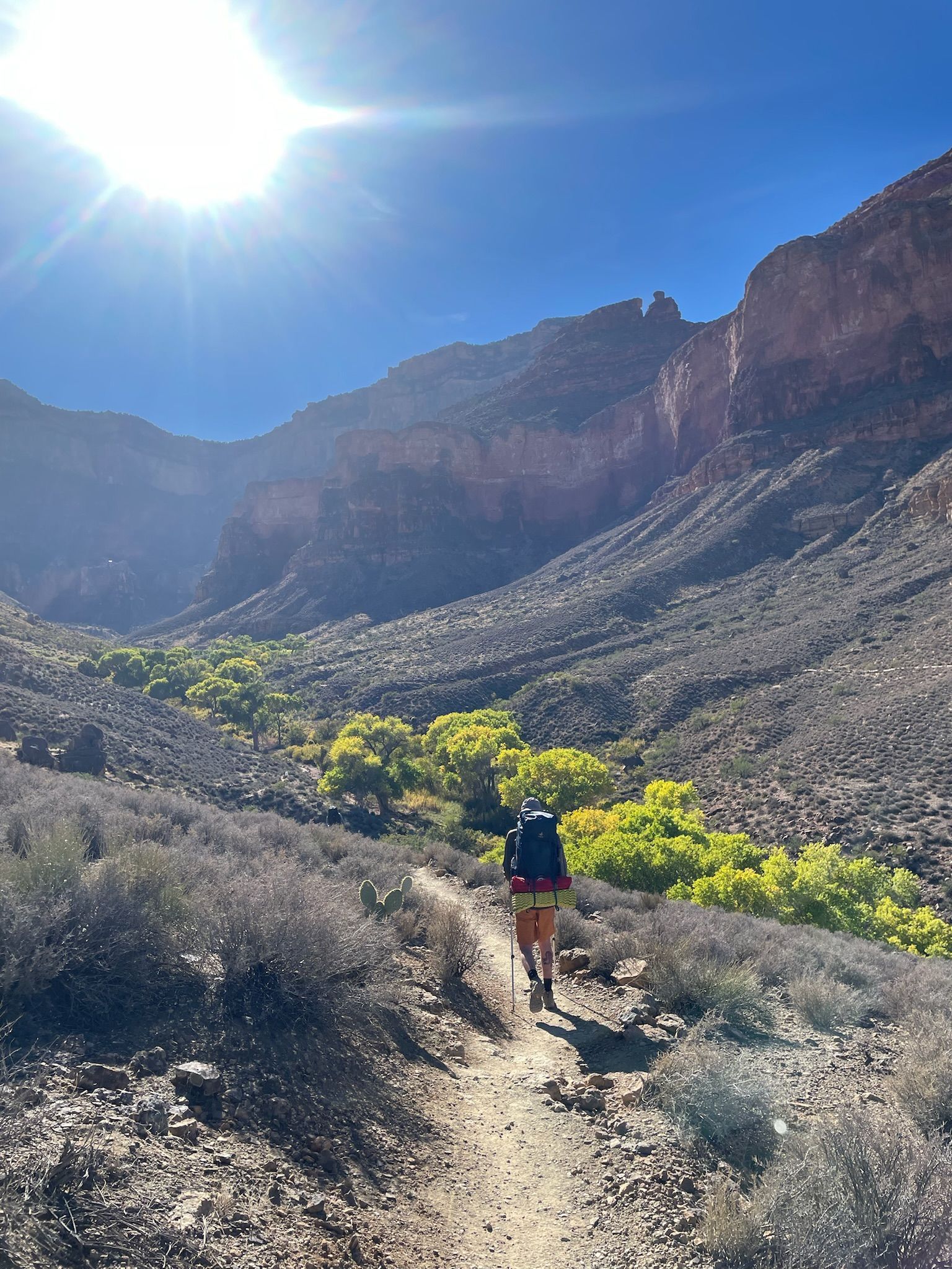 Hiker with backpack on a path in a canyon, sun shining. Green trees in the distance, dark rock walls.