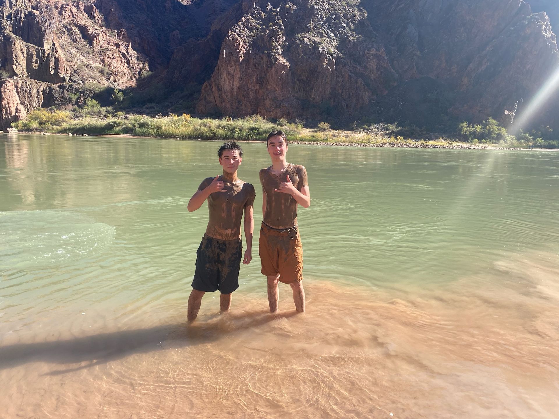Two people covered in mud stand in a river, Grand Canyon. Brown and green landscape.