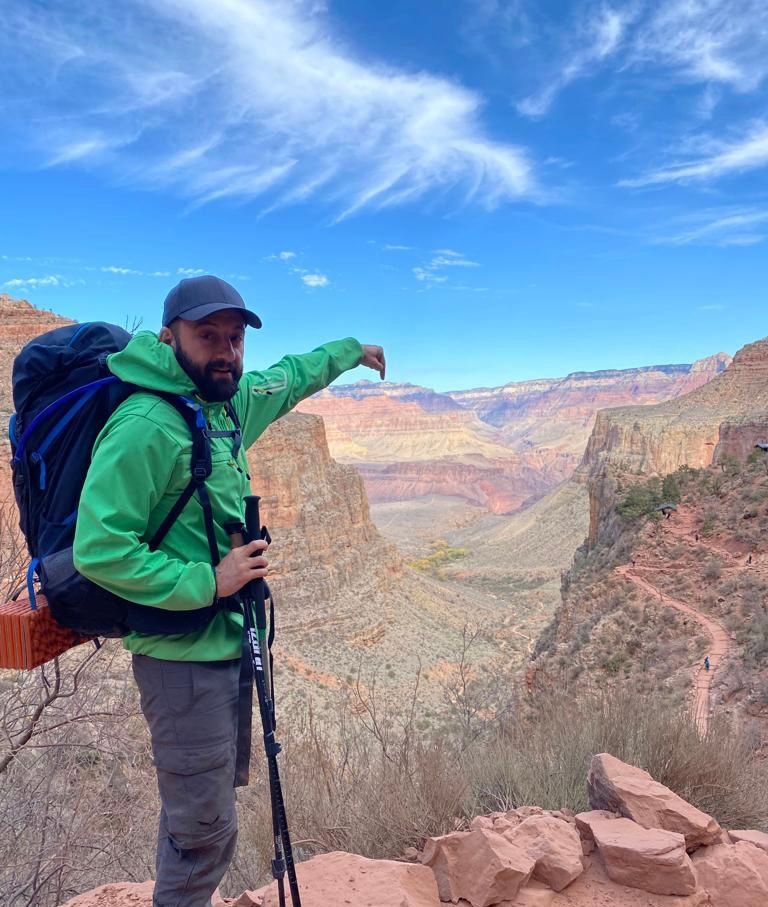 Man with backpack points at Grand Canyon vista under blue sky.