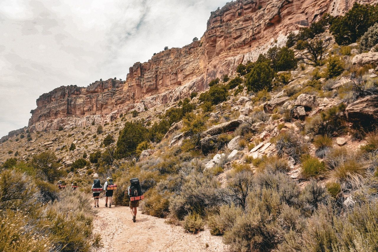 Hikers walking on a dirt trail towards a rocky cliff under a cloudy sky.