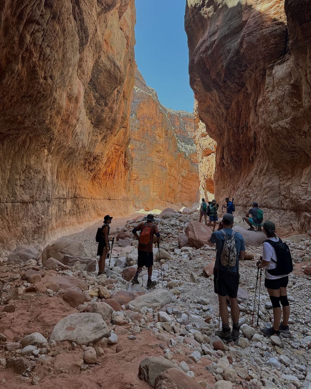 Hikers in a narrow red rock canyon, walking over rocks, under a blue sky.
