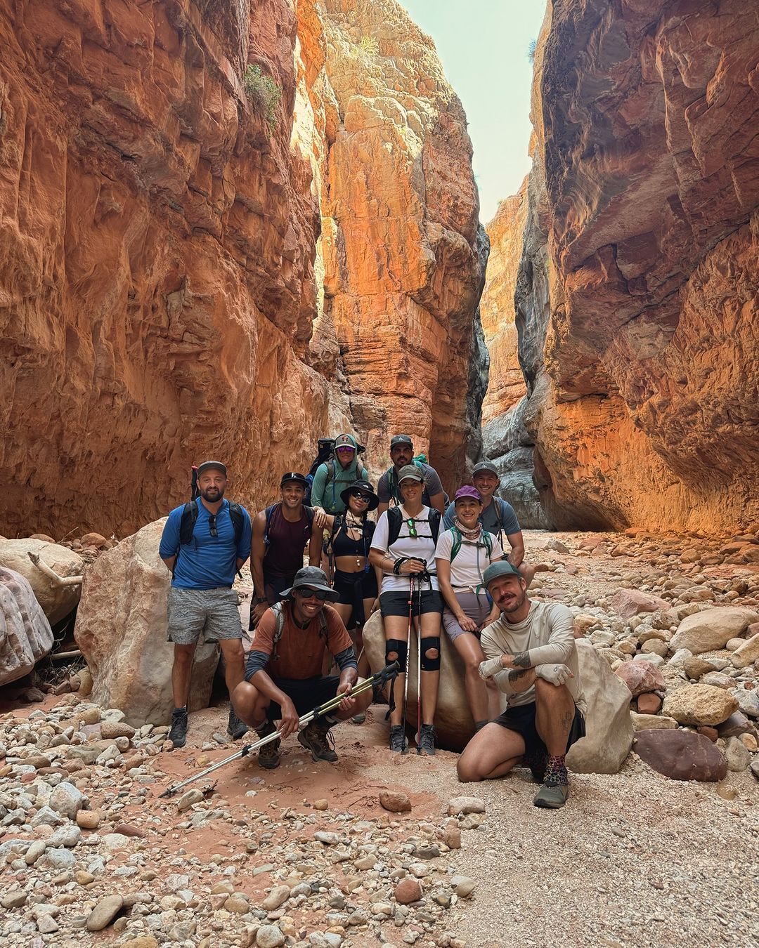 Group of people standing in a narrow, red-rock canyon. They appear to be hiking on a gravel trail. Sunny day.