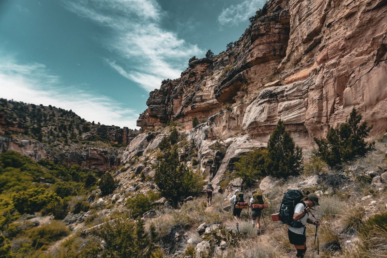 Hikers with backpacks on a trail, near a canyon wall and shrubs under a cloudy sky.