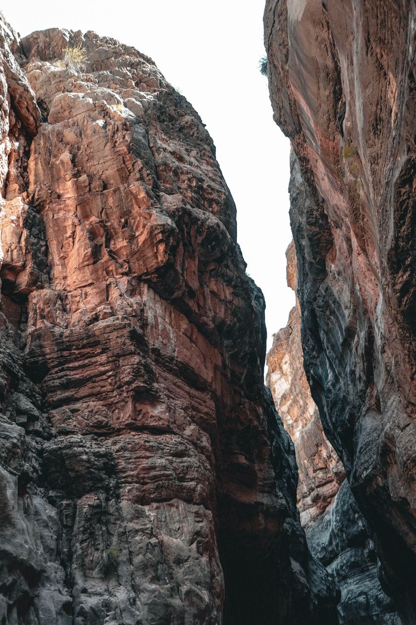 Narrow canyon walls in shades of brown and red, opening to a bright sky above.