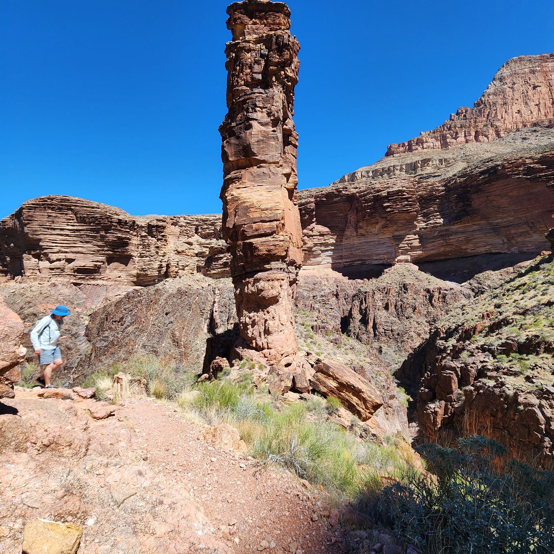 Person hiking by a tall, red rock spire in a canyon on a sunny day.