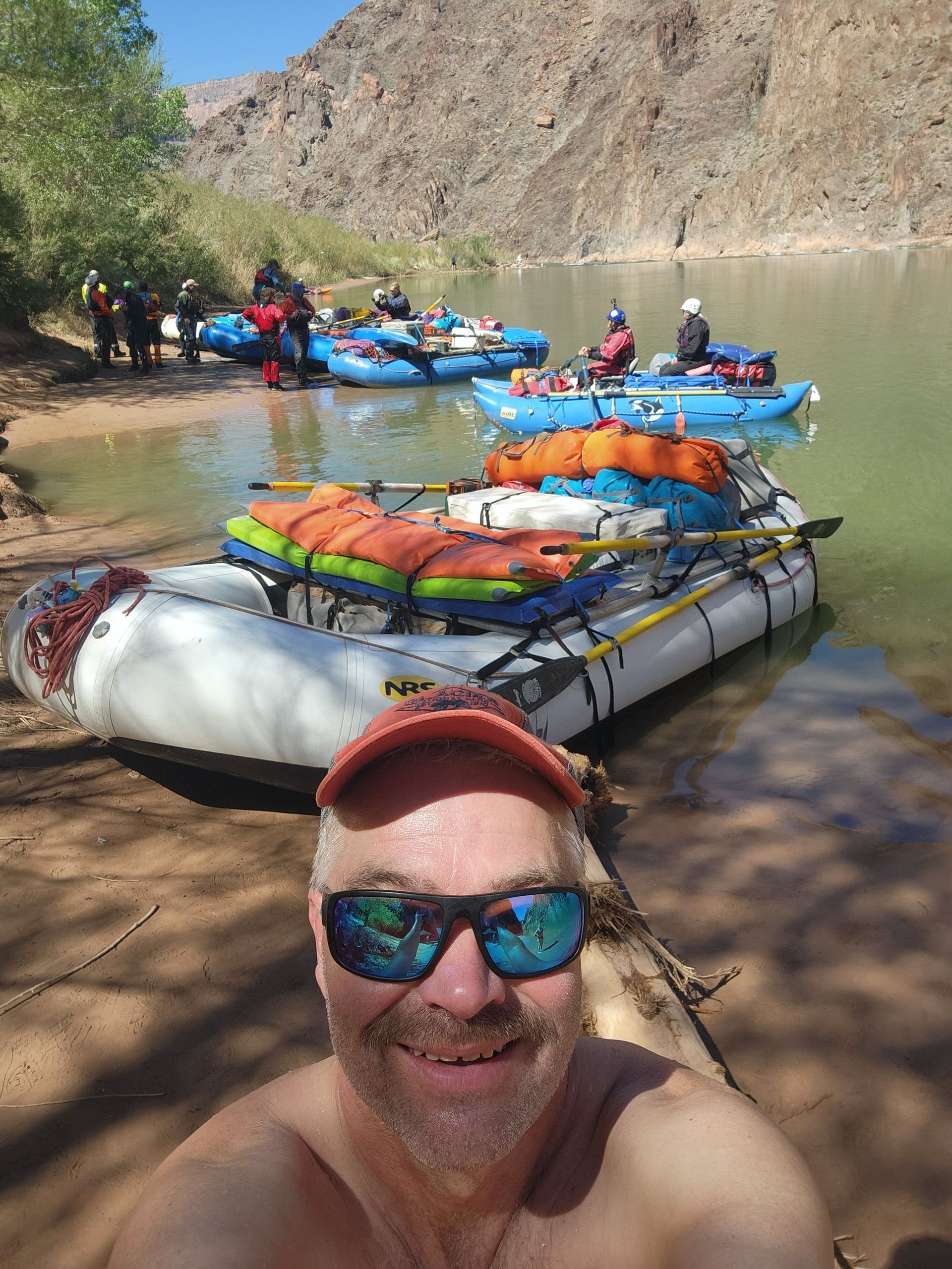 Man in sunglasses smiles at camera; rafts on shore of a river in a canyon.