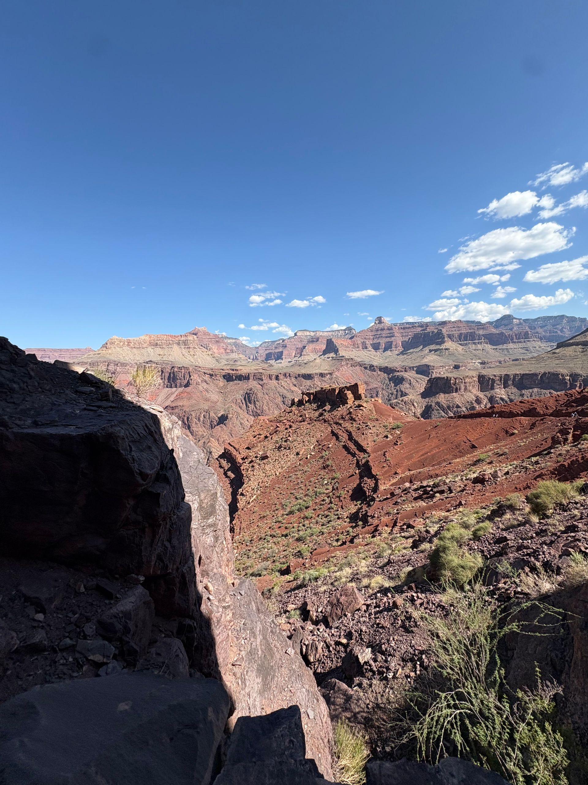 Canyon landscape: red-brown cliffs under a blue sky with sparse clouds. Rocky foreground.