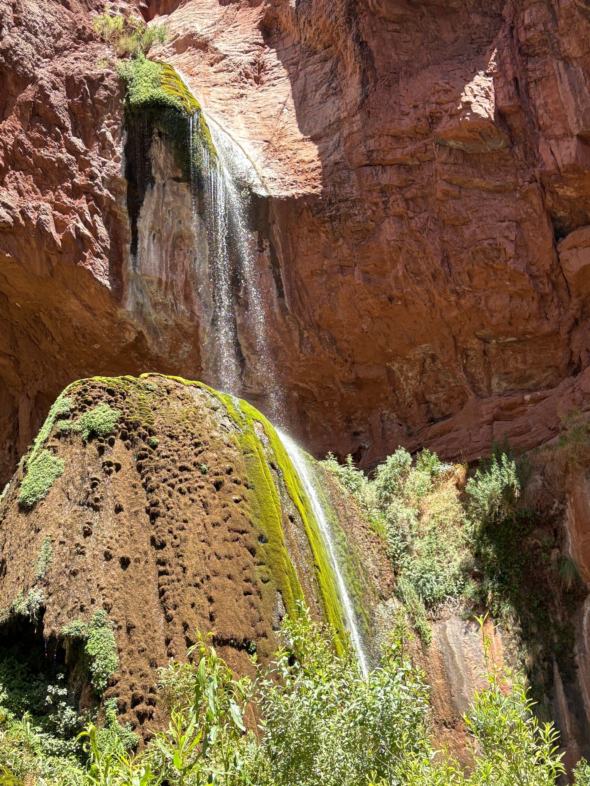 Waterfall cascading down red rock cliff face, water-covered rock covered in green vegetation.