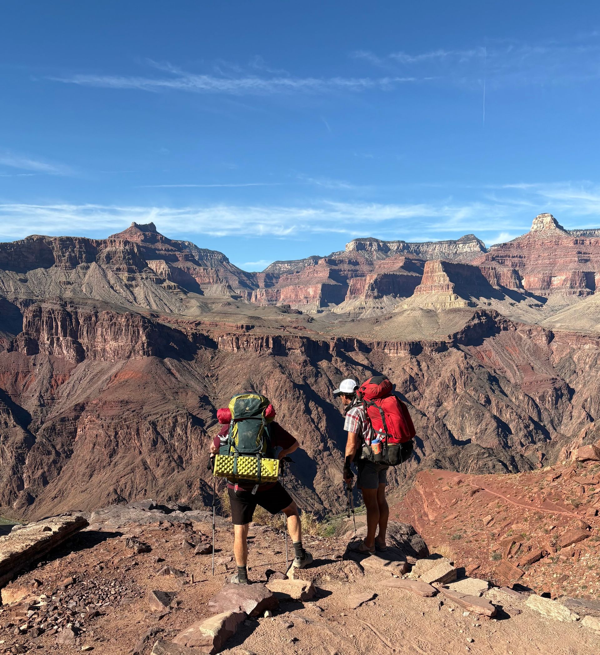 Two hikers with backpacks on a red rock trail, overlooking a canyon under a blue sky.