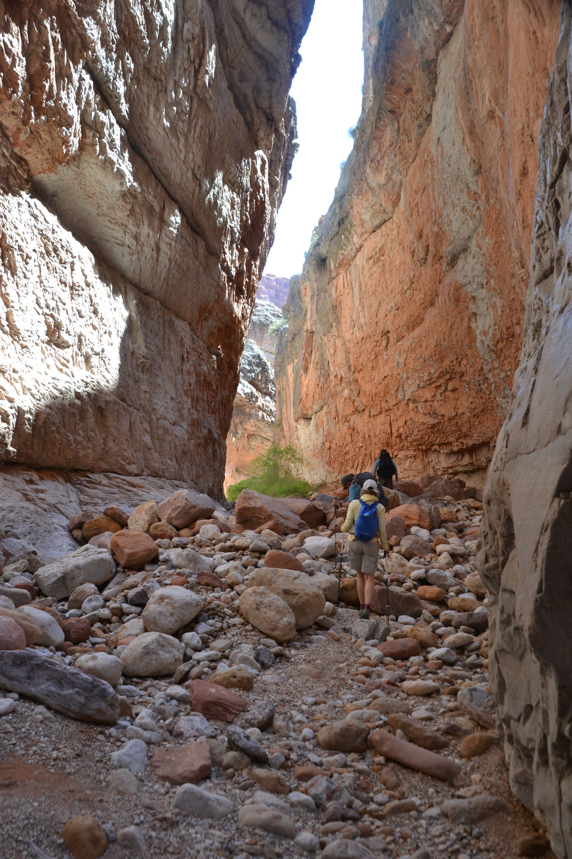 Hikers in narrow canyon with red rock walls, walking on rocky path.