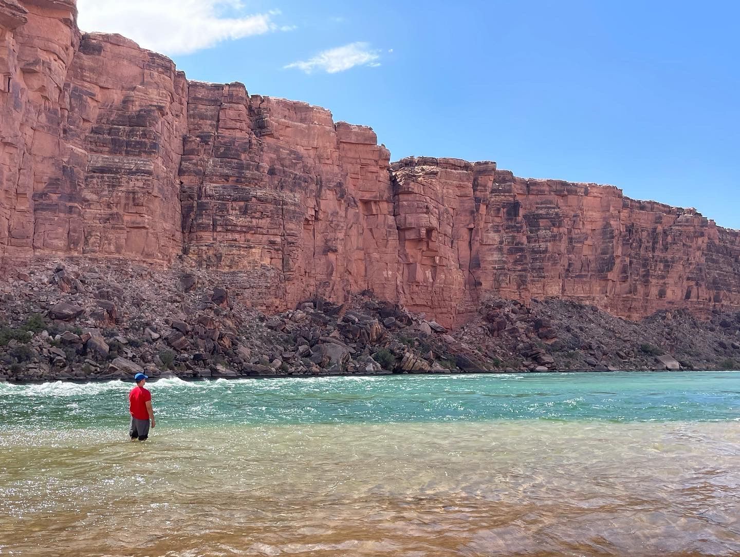 A man in a red shirt is standing in the water near a cliff.