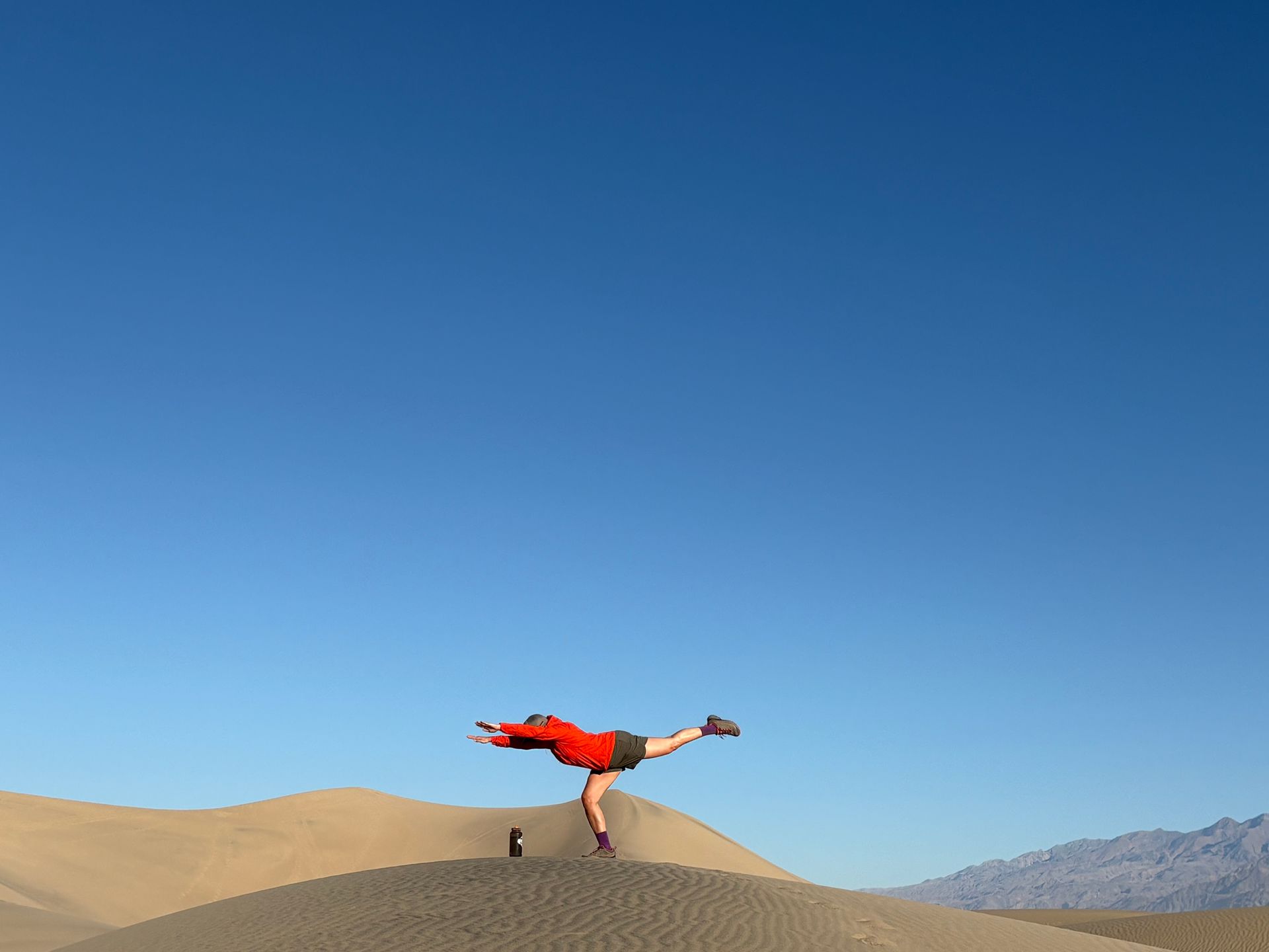 Person balancing on one leg atop a sand dune, arms outstretched, against a clear blue sky.