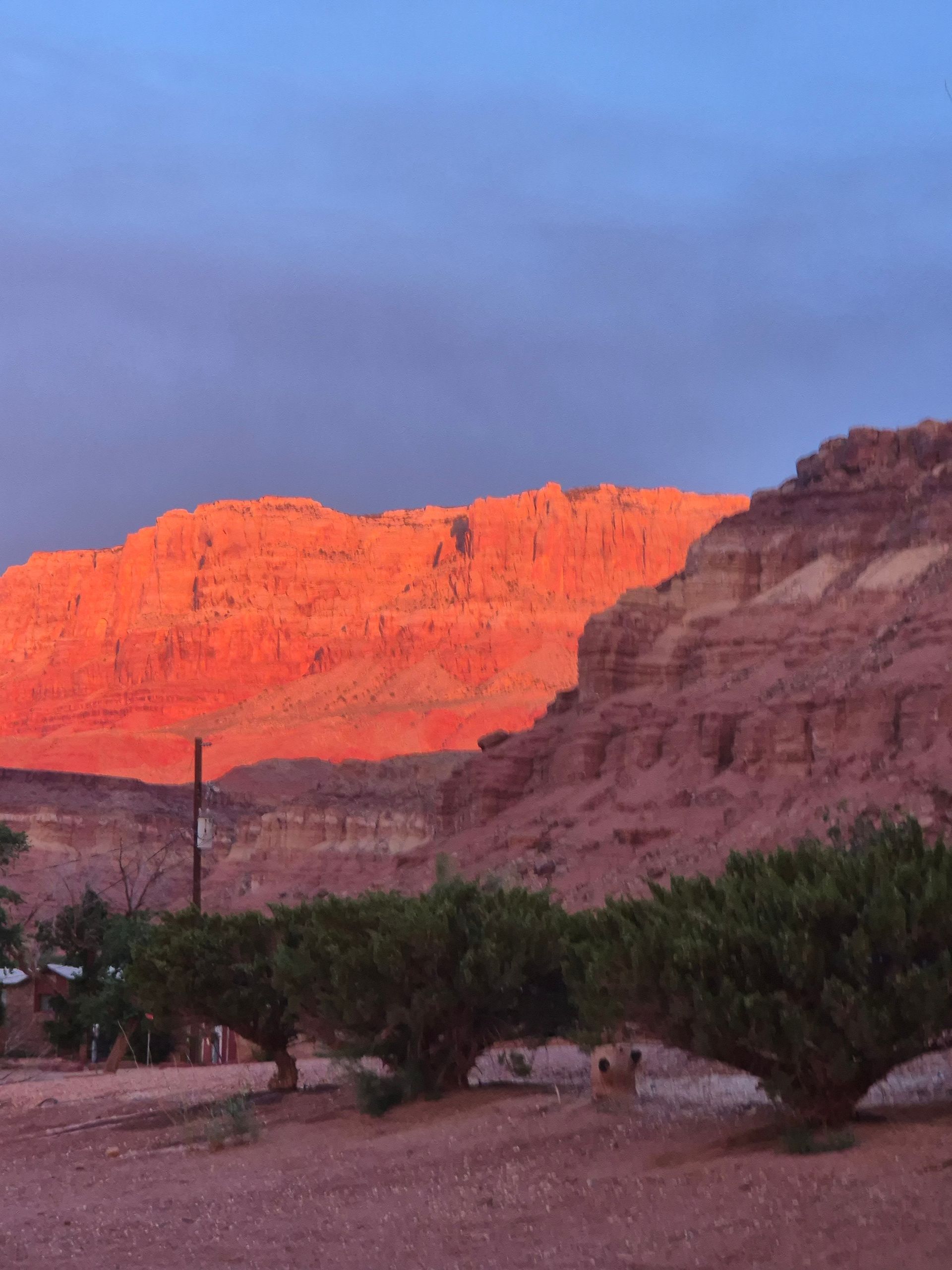 Sunset illuminates red rock cliffs with a fiery glow, blue sky above, green shrubs below.