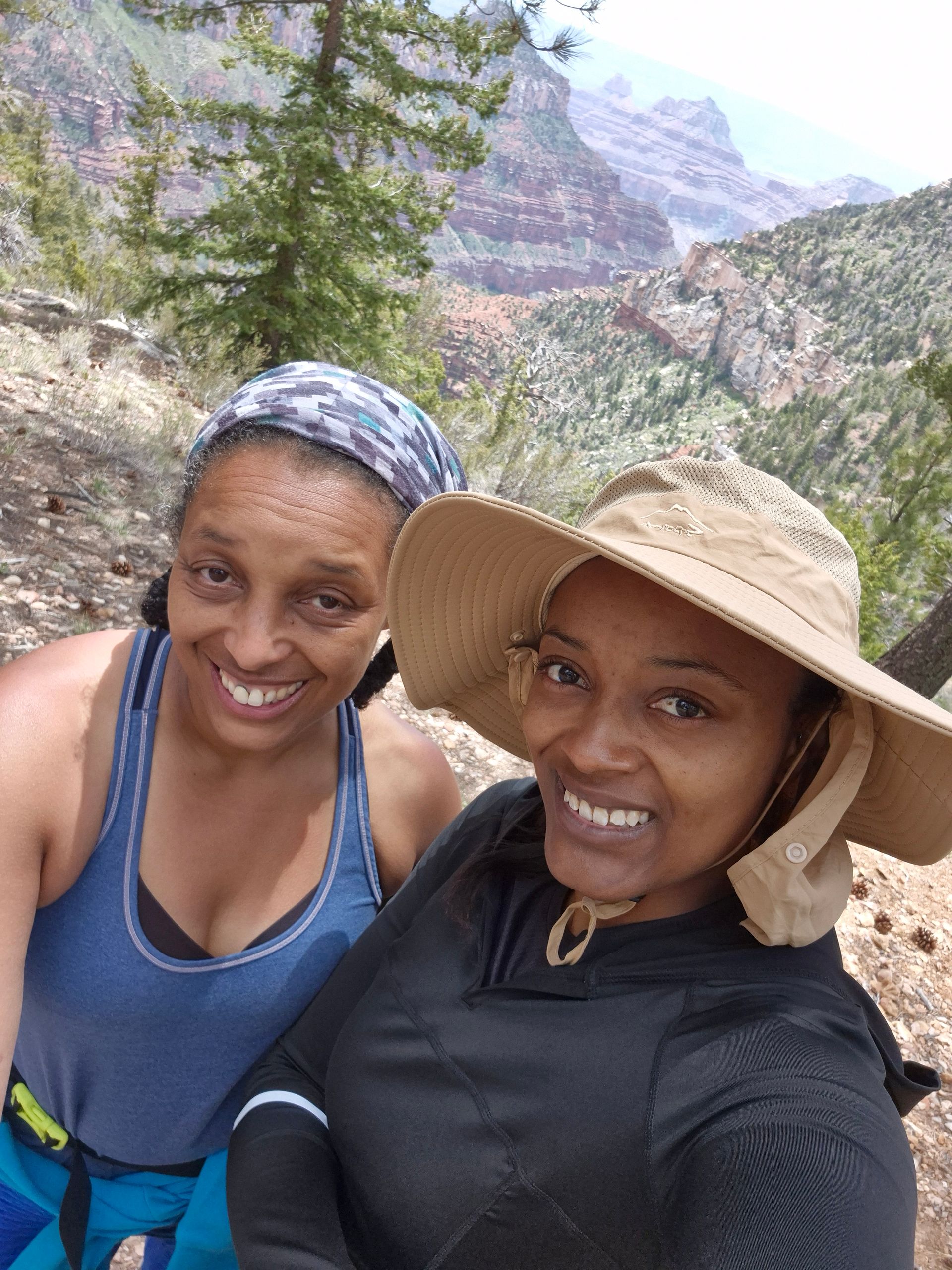 Two women smile, posing outdoors with mountains in the background. One wears a hat, the other a headband.