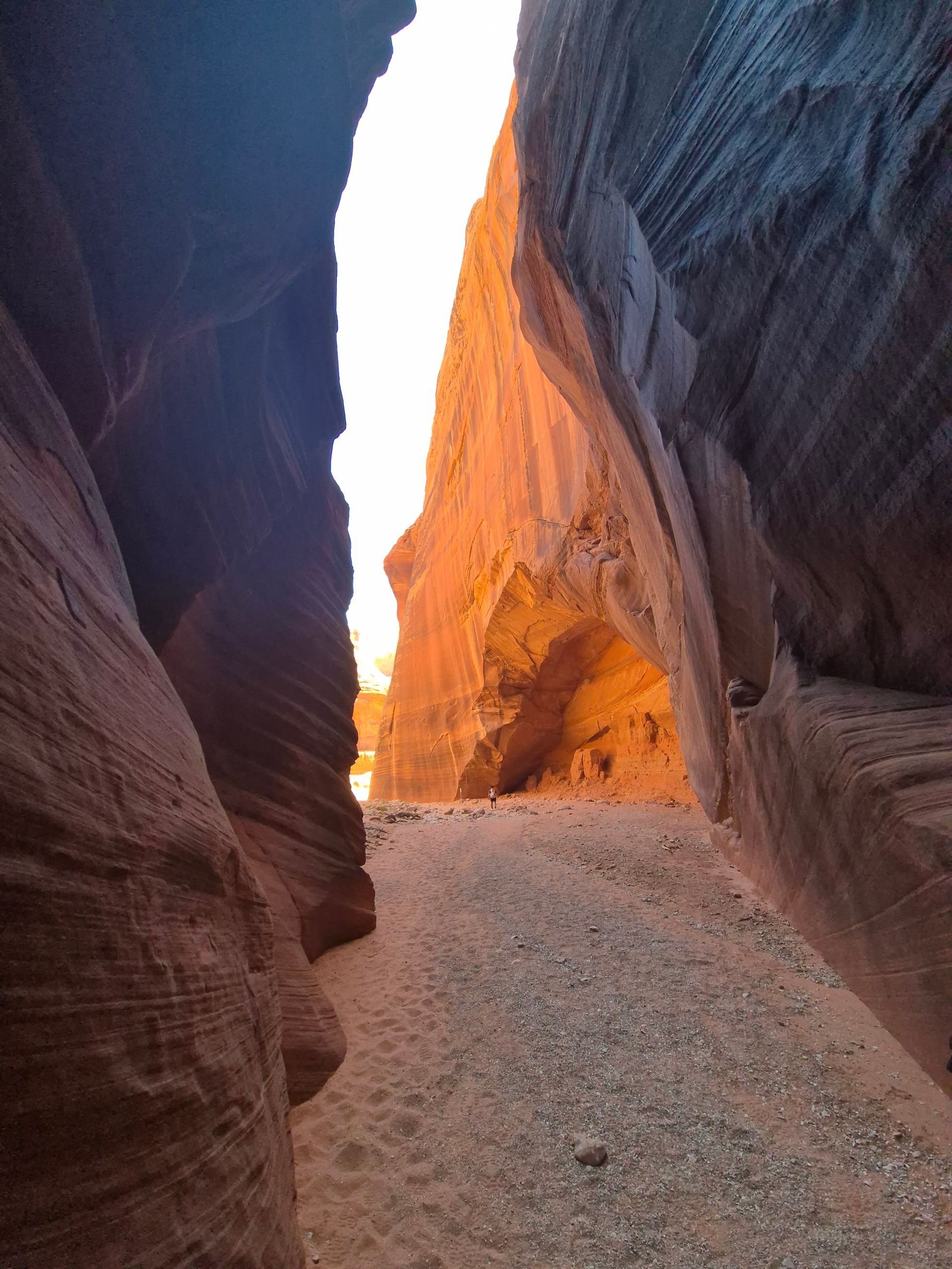 Narrow sandstone canyon, light at end. Red-orange walls, gray ground, sunlight.