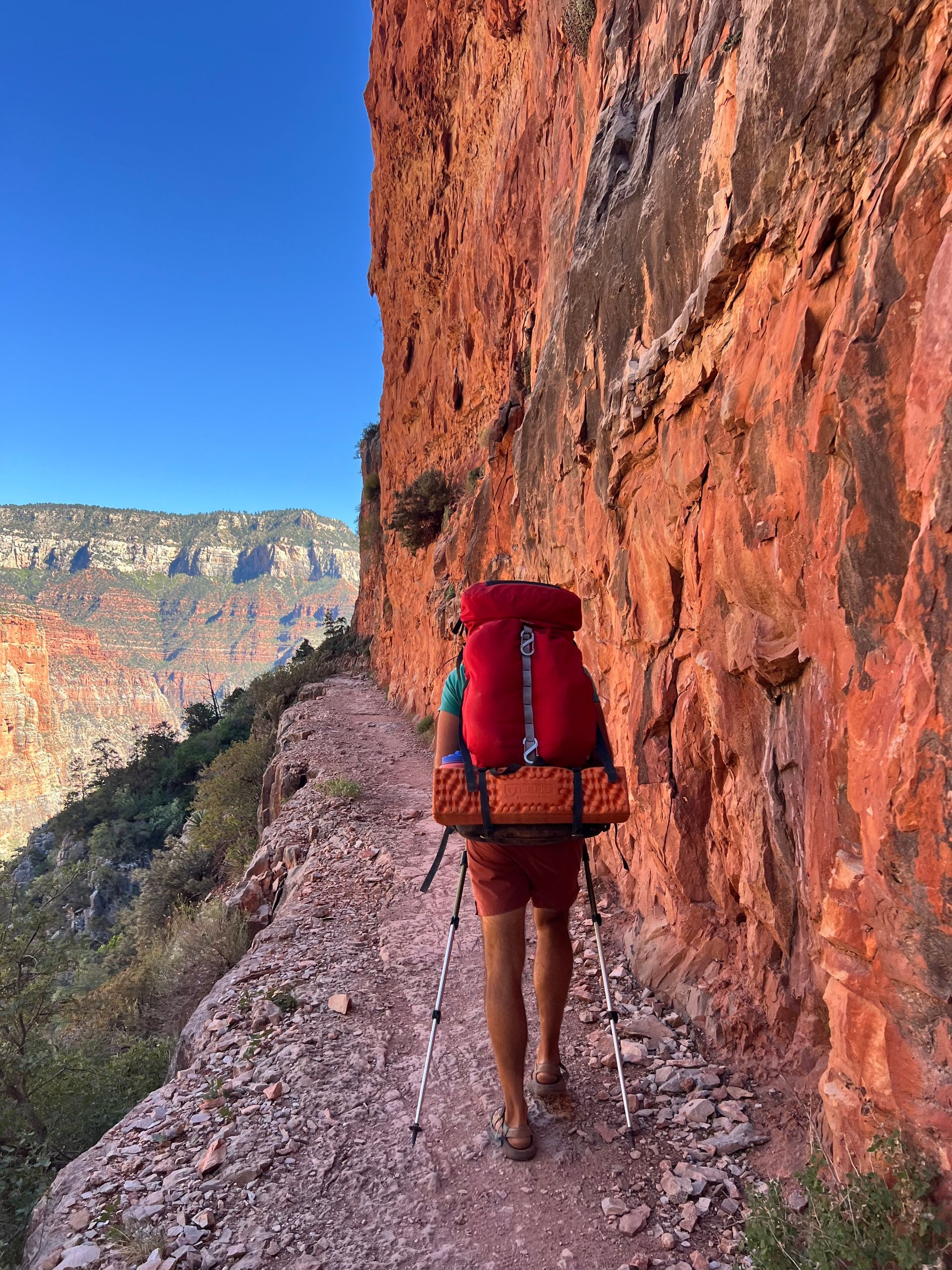 Person hikes narrow trail along red canyon wall. Backpack, blue sky, and canyon view.