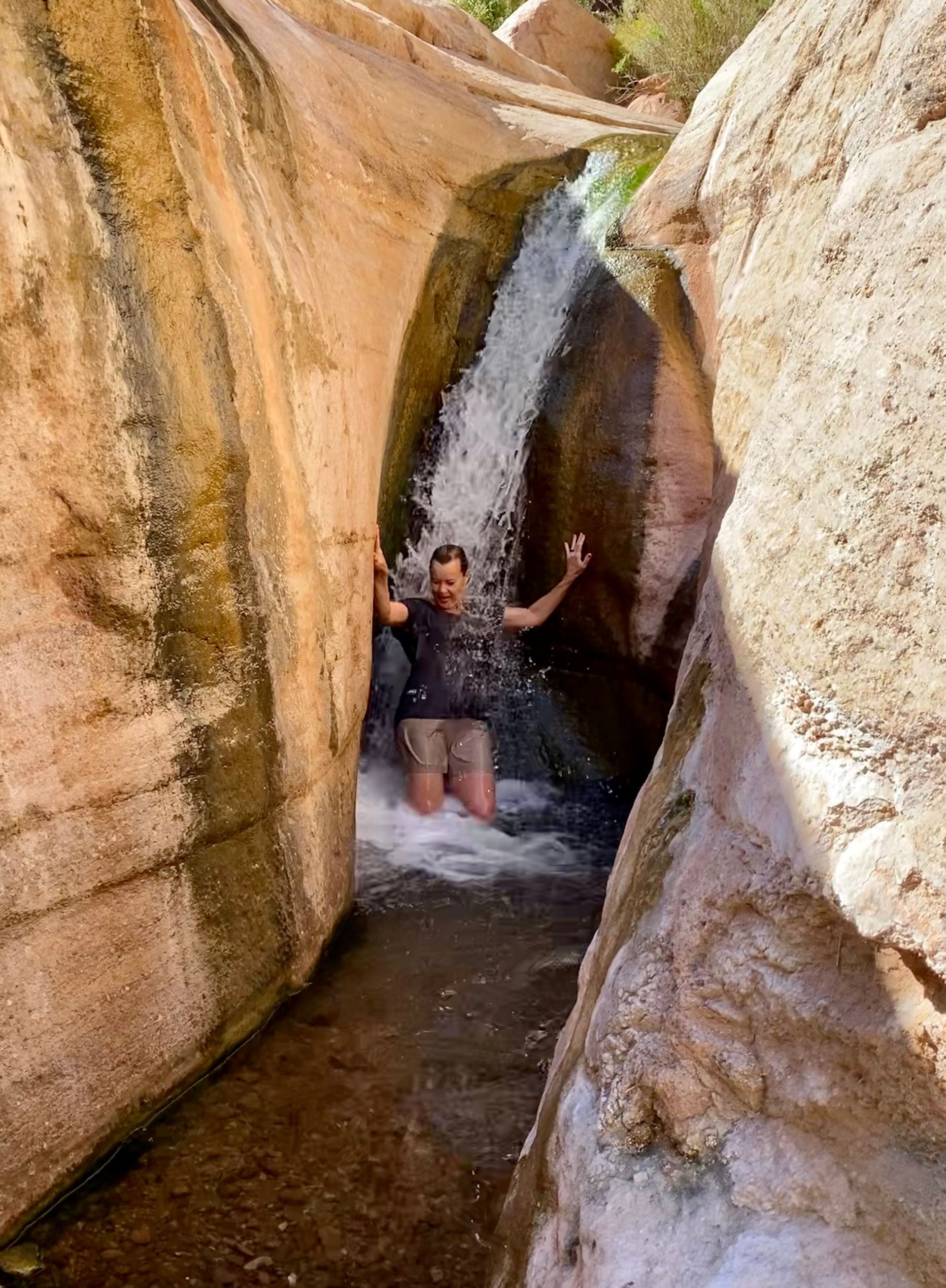 A man is kneeling in front of a waterfall in a canyon.