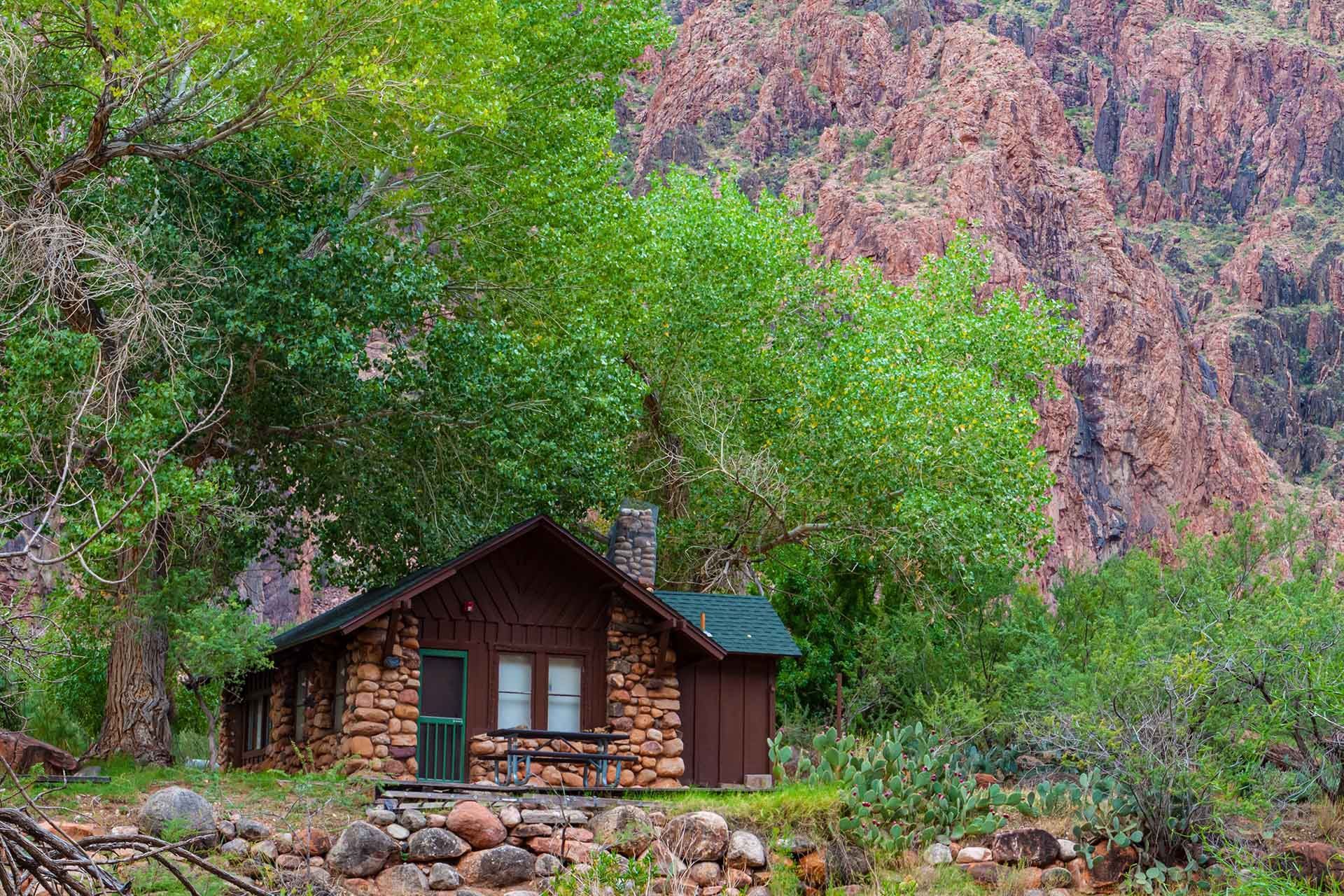 A small log cabin is surrounded by trees and rocks in the middle of a forest.