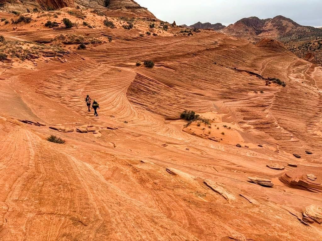 Two hikers trek across reddish-orange sandstone landscape. Cloudy sky overhead.