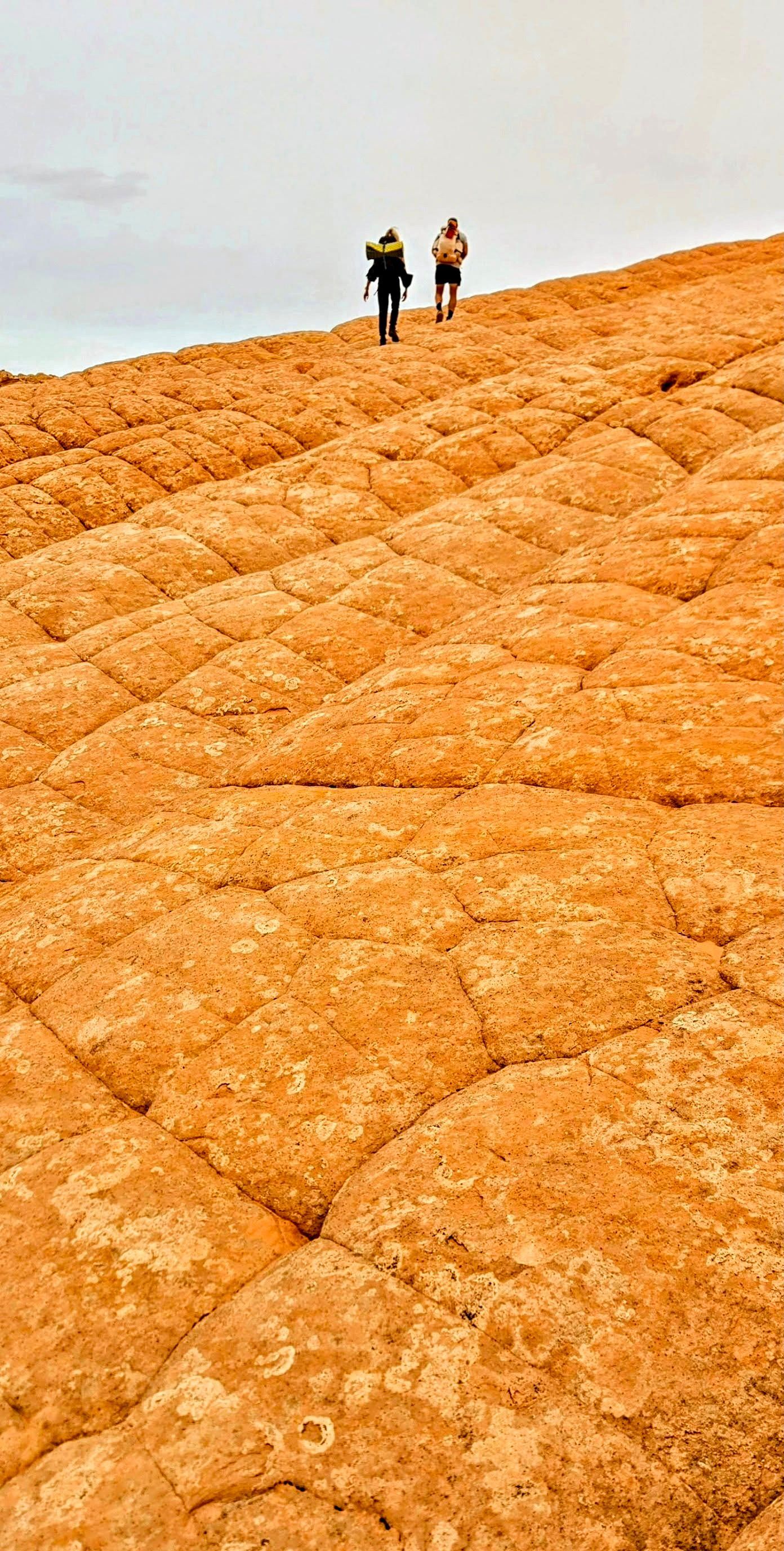 Two people hike up a textured, orange rock formation under an overcast sky.