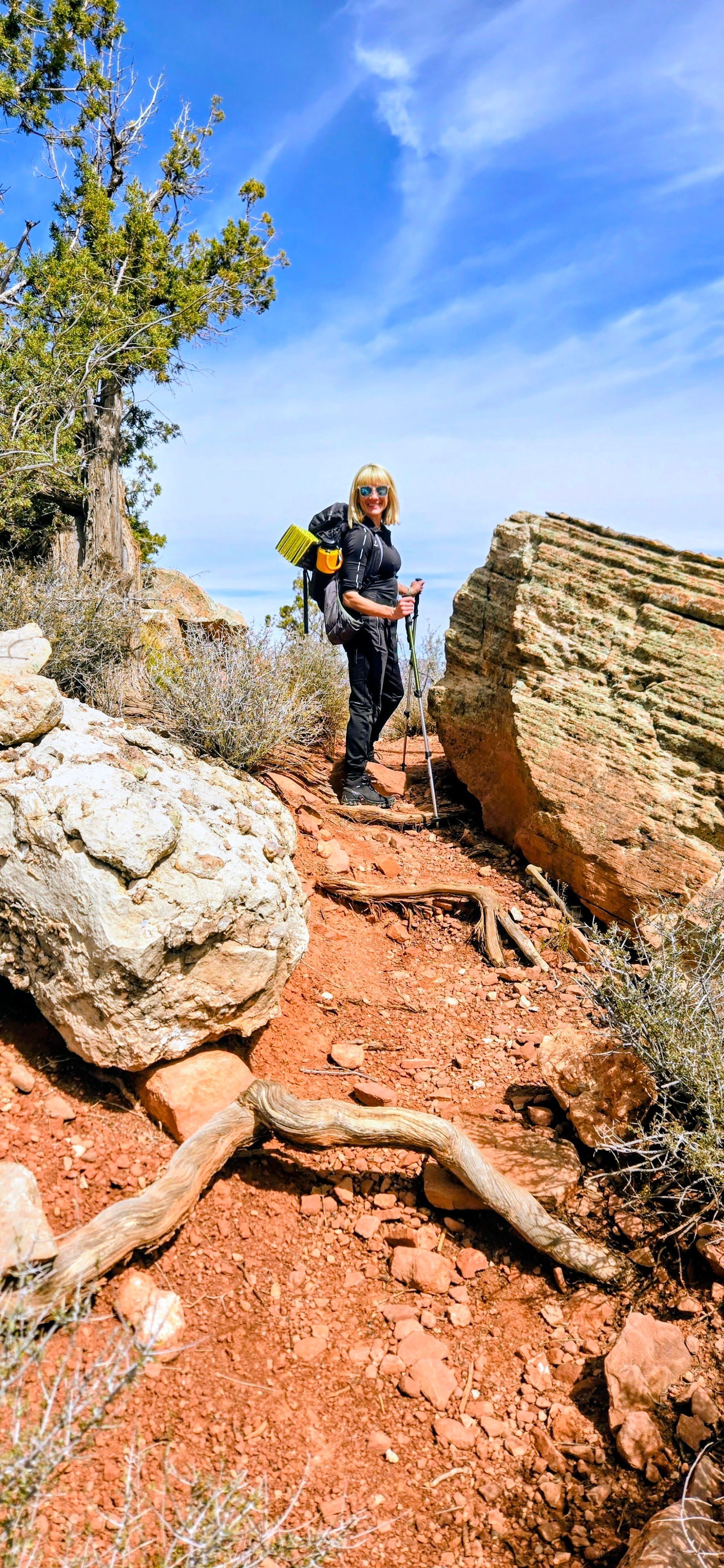 Hiker with backpack and trekking poles on a red rock trail under a blue sky.