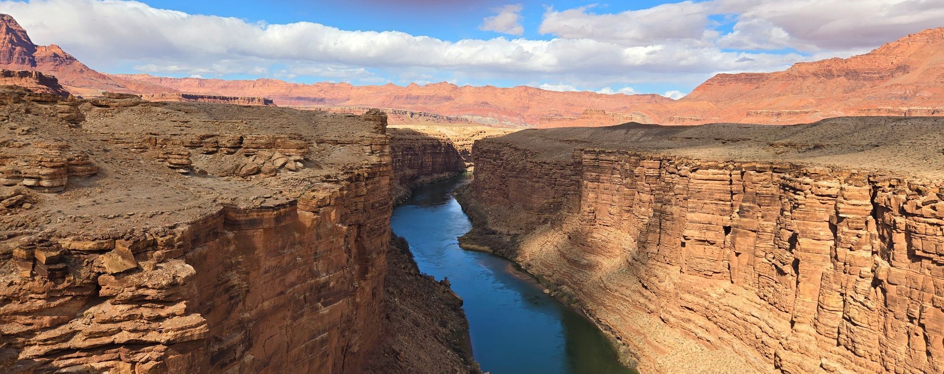 A canyon with a river flowing through it. The canyon walls are reddish-brown, and the sky is blue with clouds.