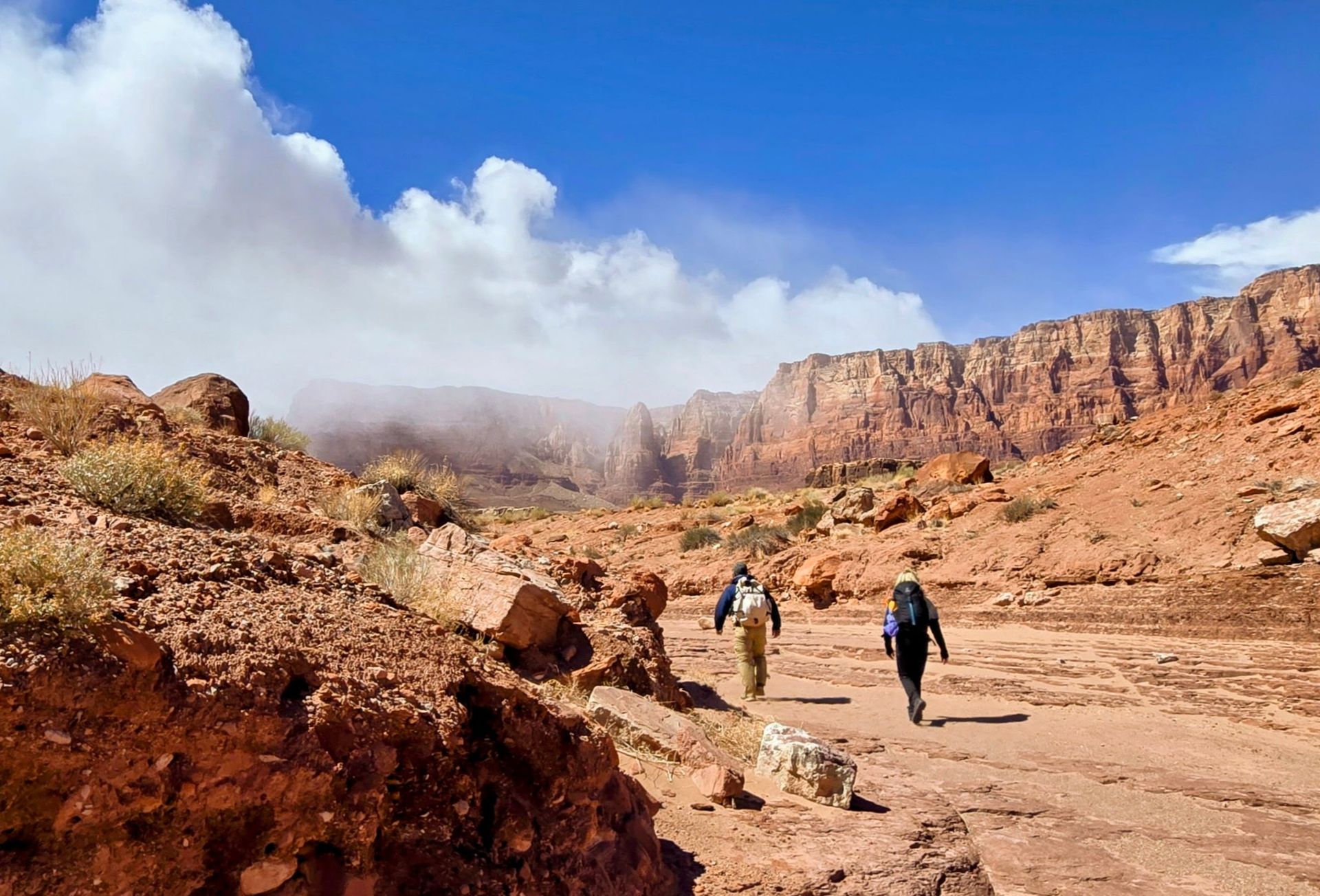 Hikers on a dusty trail in a red rock canyon under a blue sky with clouds.