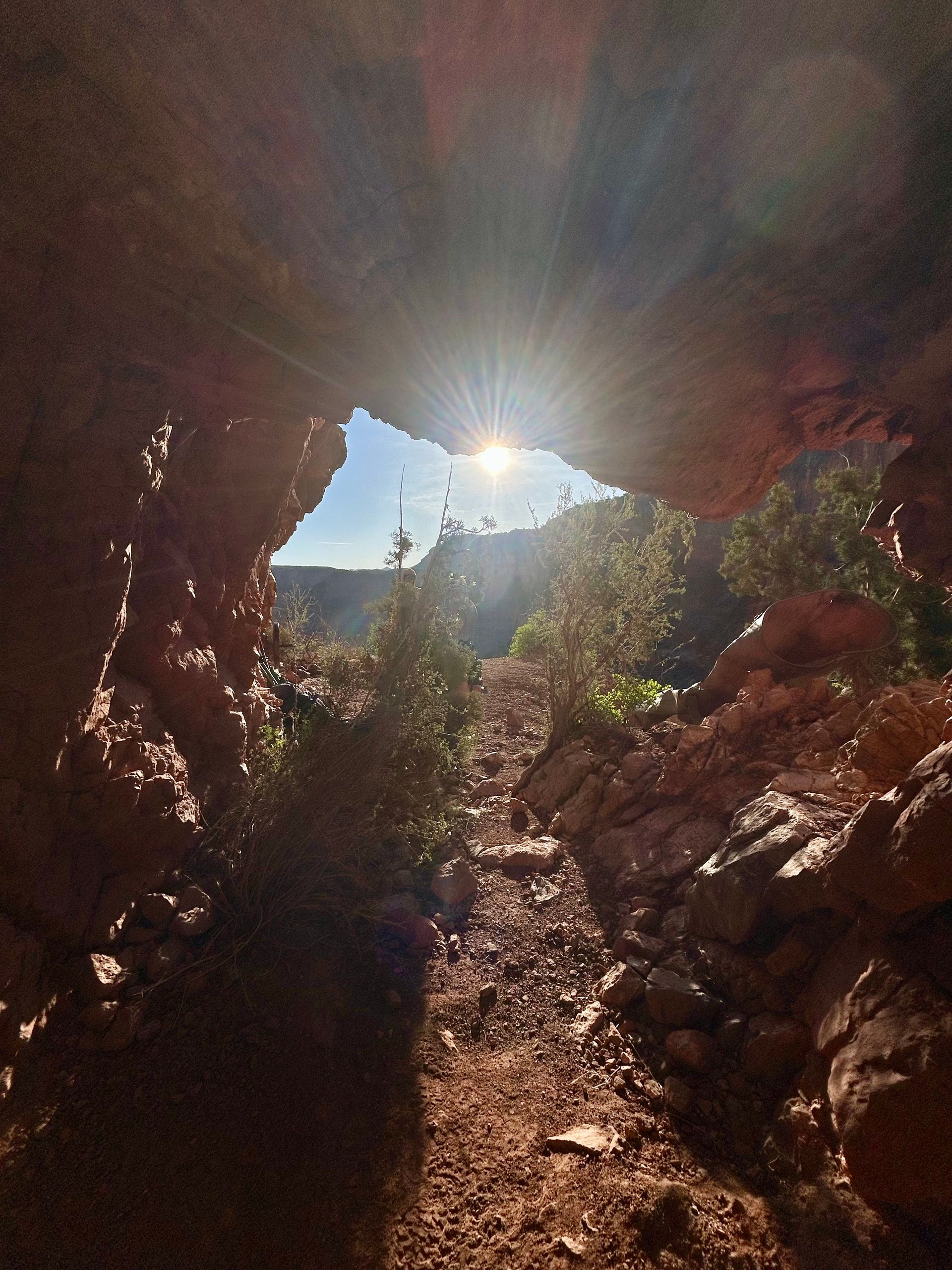 Sunlight streams through a cave opening onto a dirt path surrounded by reddish rock and vegetation.