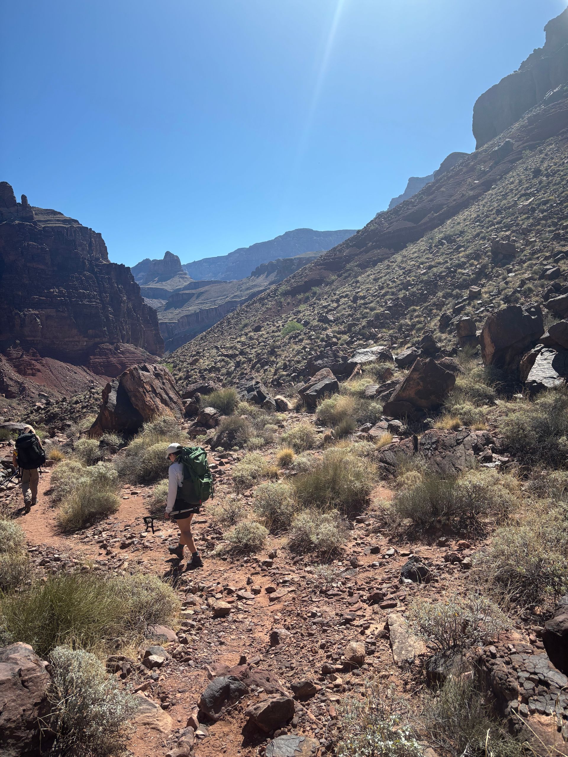 Hikers with backpacks traverse a rocky canyon trail under a bright blue sky.