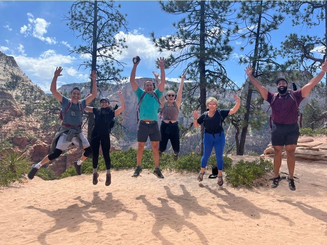Group of people jumping joyfully on a trail in a sunny, scenic mountain landscape.